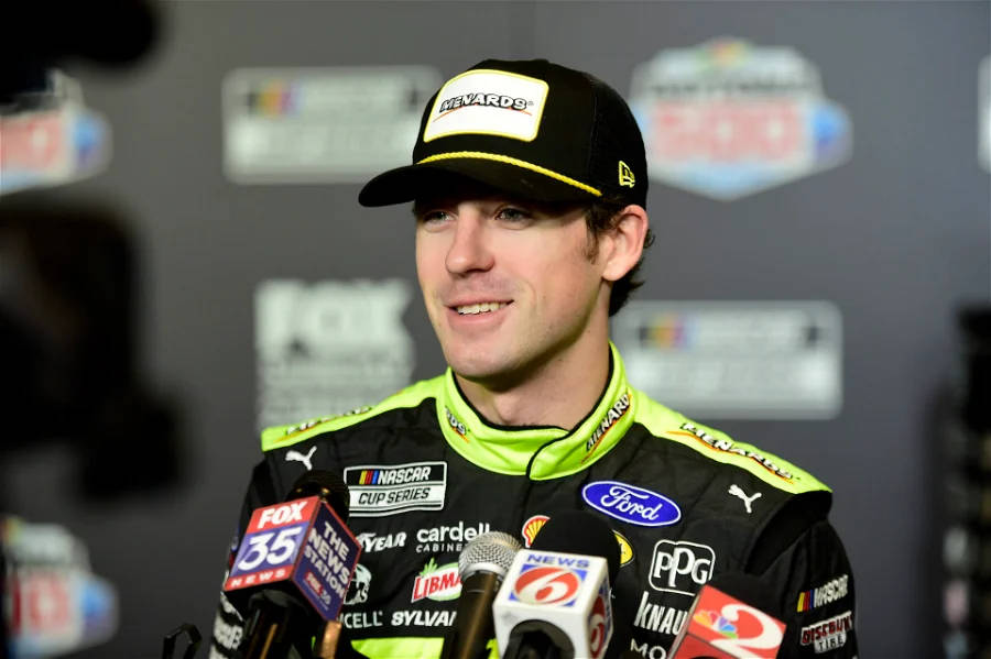 Ryan Blaney 桌布 壁紙 — NASCAR車手在記者會上自信地微笑，周圍環繞著麥克風和贊助商標誌。