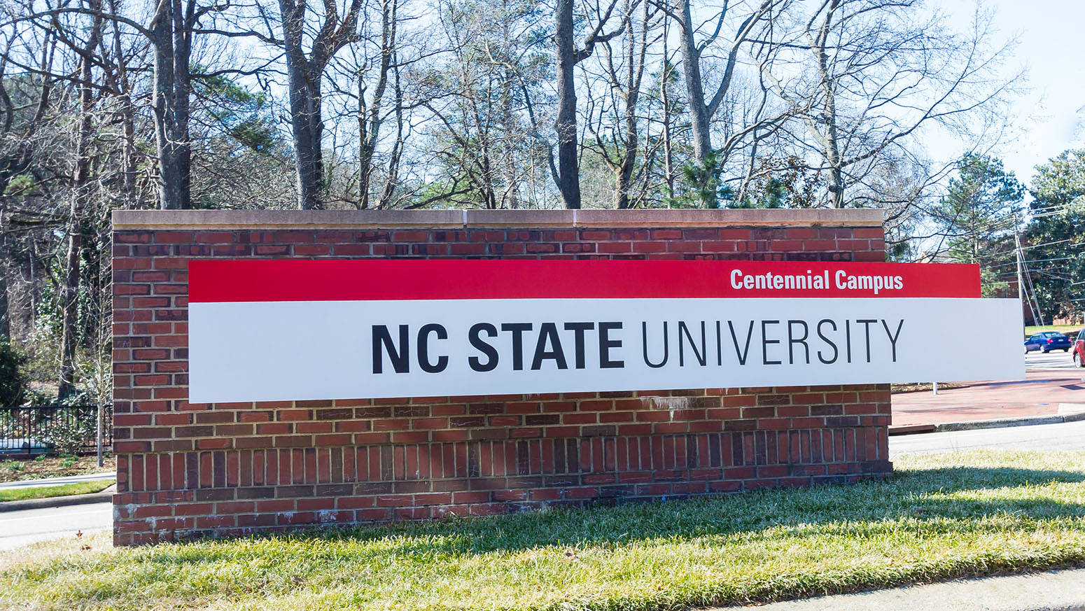 Brick Wall 桌布 壁紙 — NC State University Centennial Campus 的磚製標誌矗立在草地上，背後有光禿的樹木，在明亮的日光下。