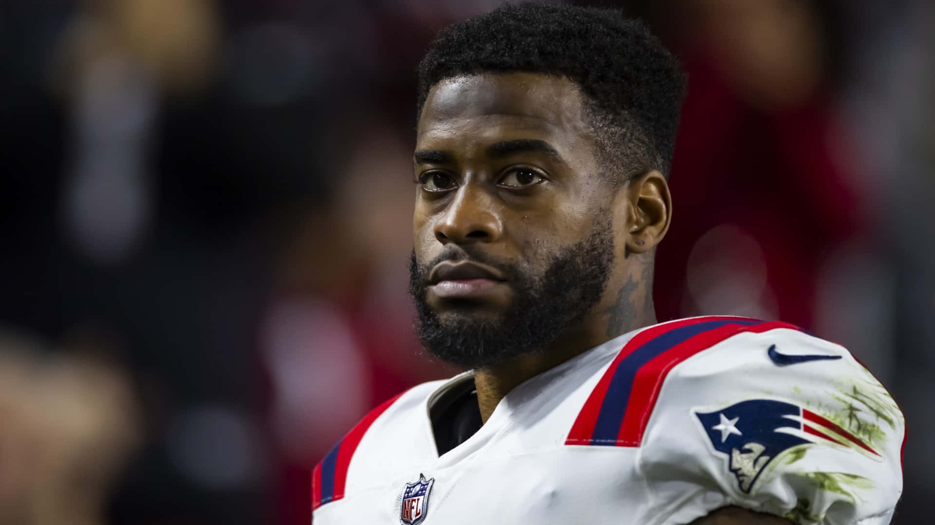  壁紙 — 一位專注的NFL球員身穿新英格蘭愛國者隊球衣，在球場燈光下比賽中凝視著。