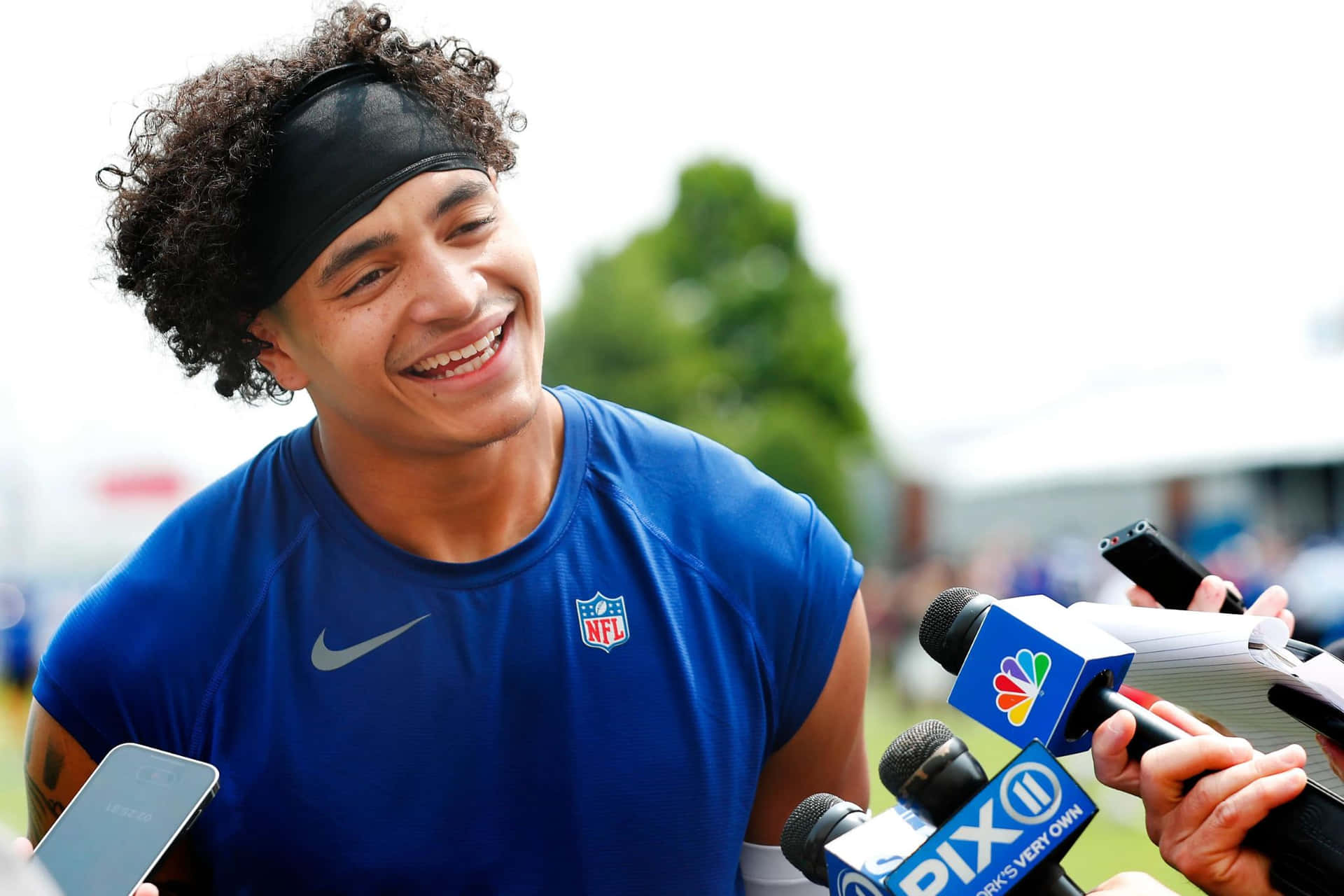  壁紙 — 一位身穿藍色球衣的NFL球員在記者會上微笑大笑，被記者和麥克風包圍。