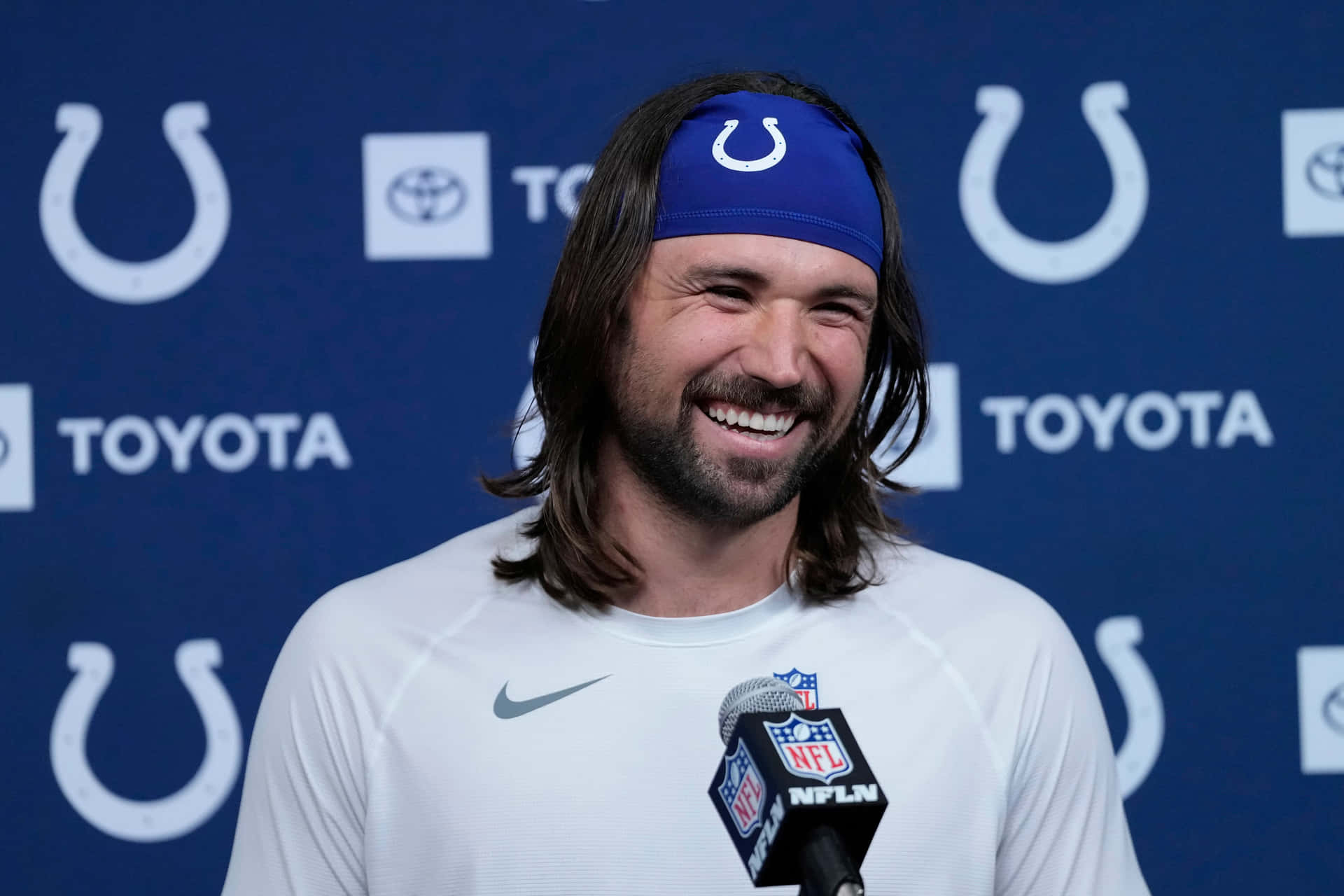 壁紙 — 一位面帶微笑的NFL球員，長髮，戴著藍色球隊頭帶，在新聞發布會上對著麥克風講話。