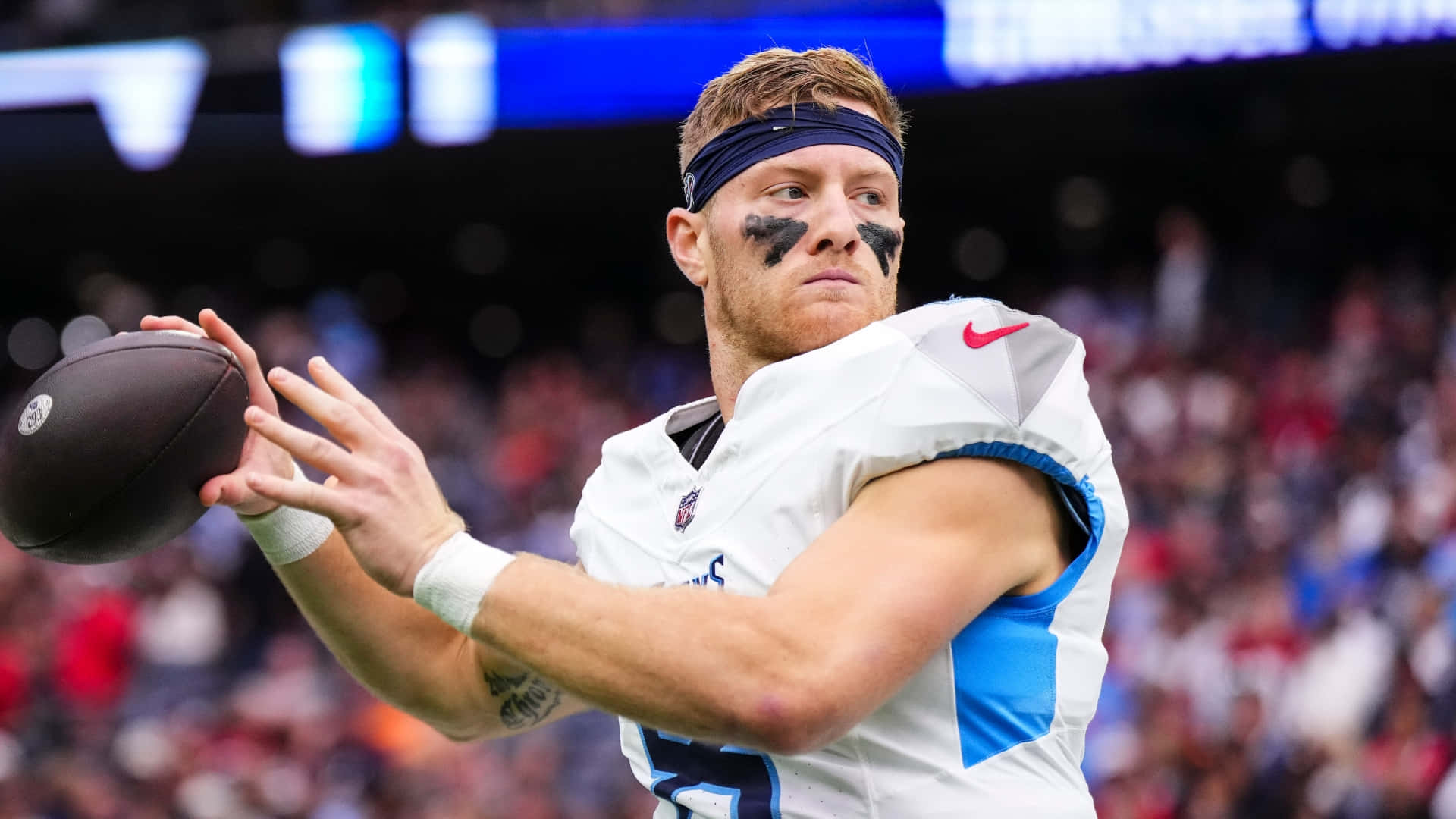  壁紙 — 一位專注的NFL四分衛身穿白色和藍色球衣，在日間比賽中準備擲出橄欖球，背景是模糊的體育場人群。