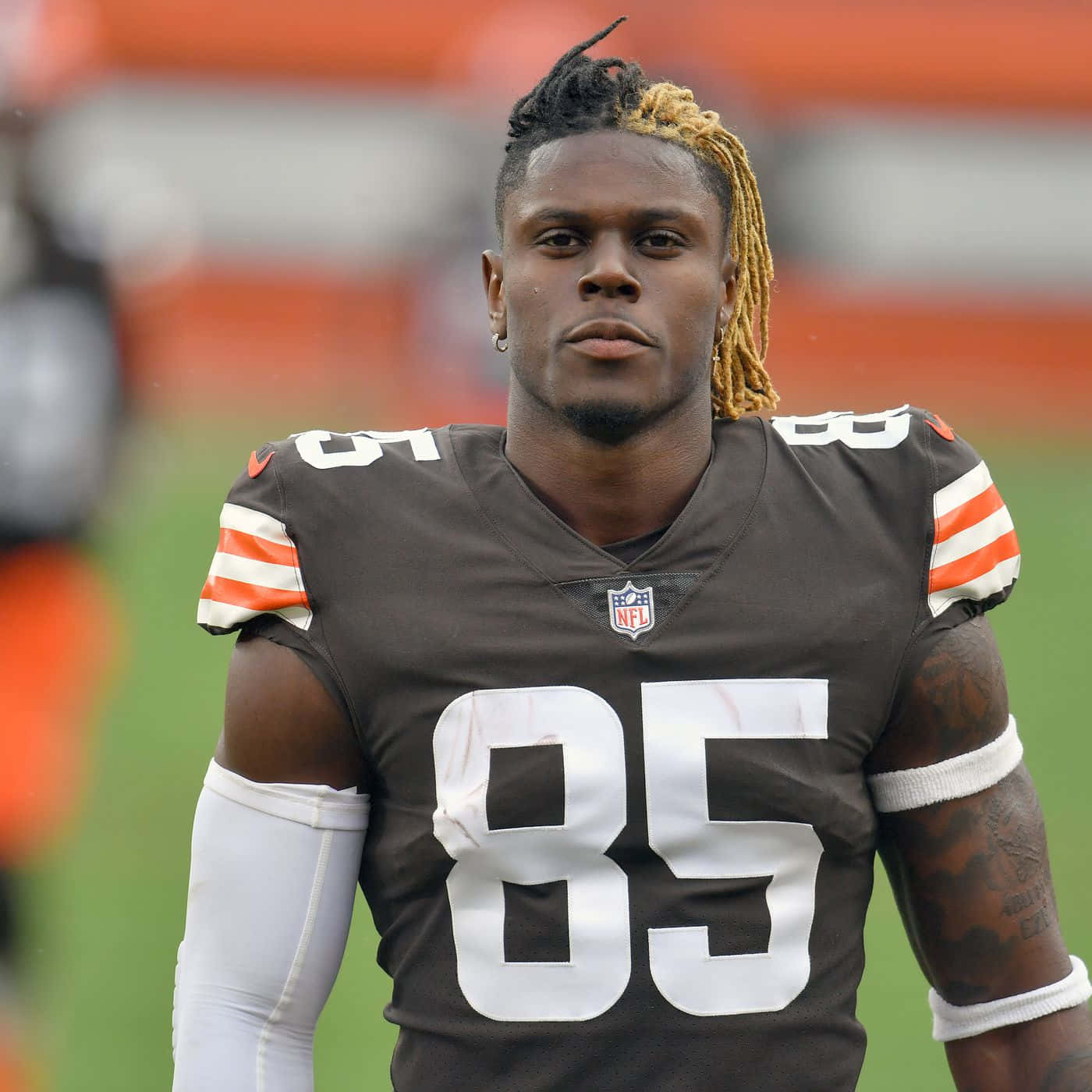  壁紙 — 一位專注的NFL外接手，身穿棕色85號球衣，站在綠色球場上，背景中橙色體育場座位模糊不清。