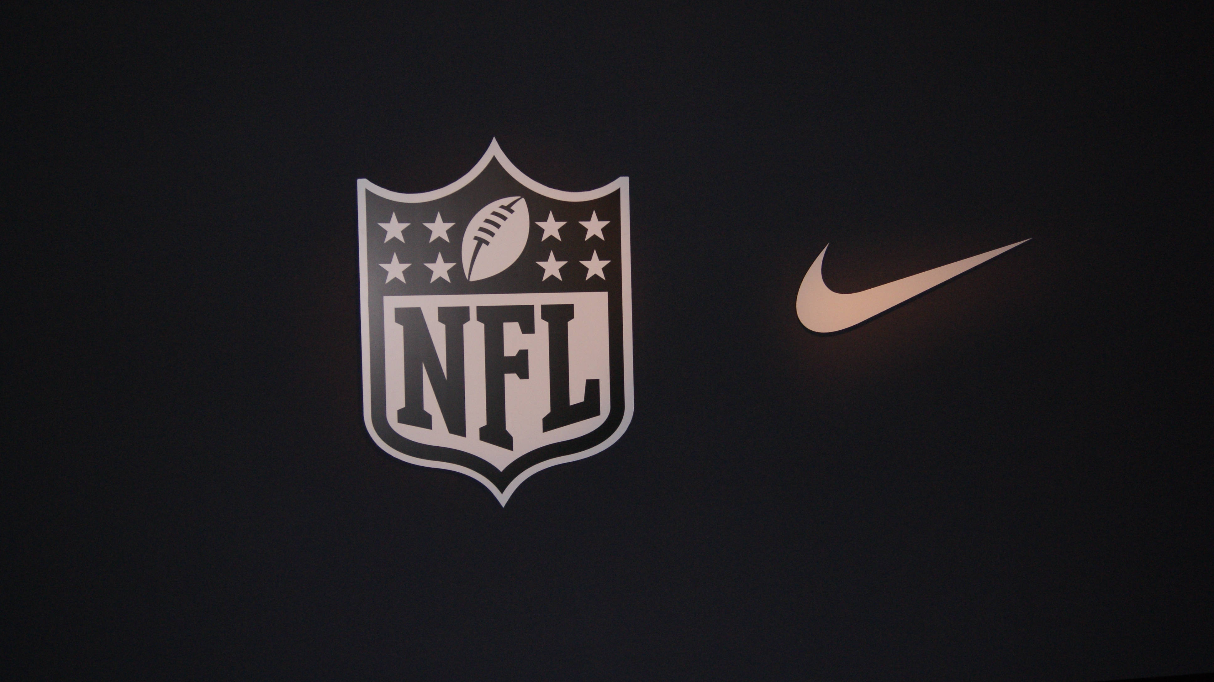 酷炫足球 桌布 壁紙 — NFL 標誌和 Nike 勾勾在深色紋理背景上柔和發光。