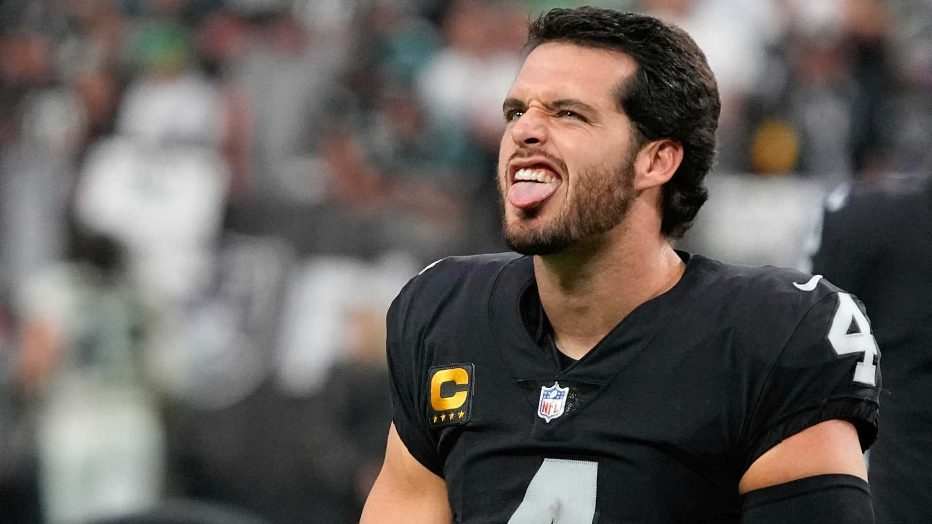 突襲者 桌布 壁紙 — NFL四分衛在比賽中伸出舌頭，表情專注，身穿黑色球衣，號碼4，帶有隊長臂章。