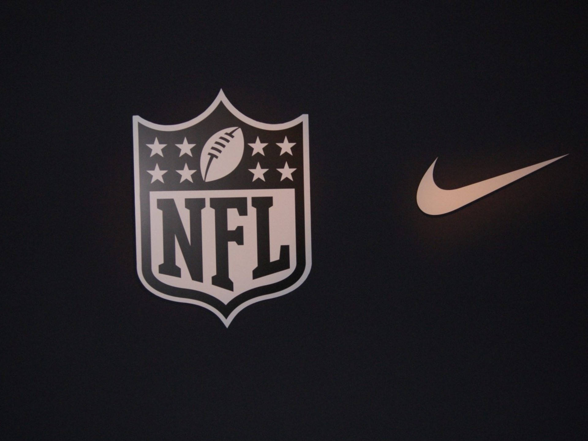 NFL 美式足球 桌布 壁紙 — NFL盾牌標誌和Nike勾勾以白色和金色呈現於深色布料背景上。