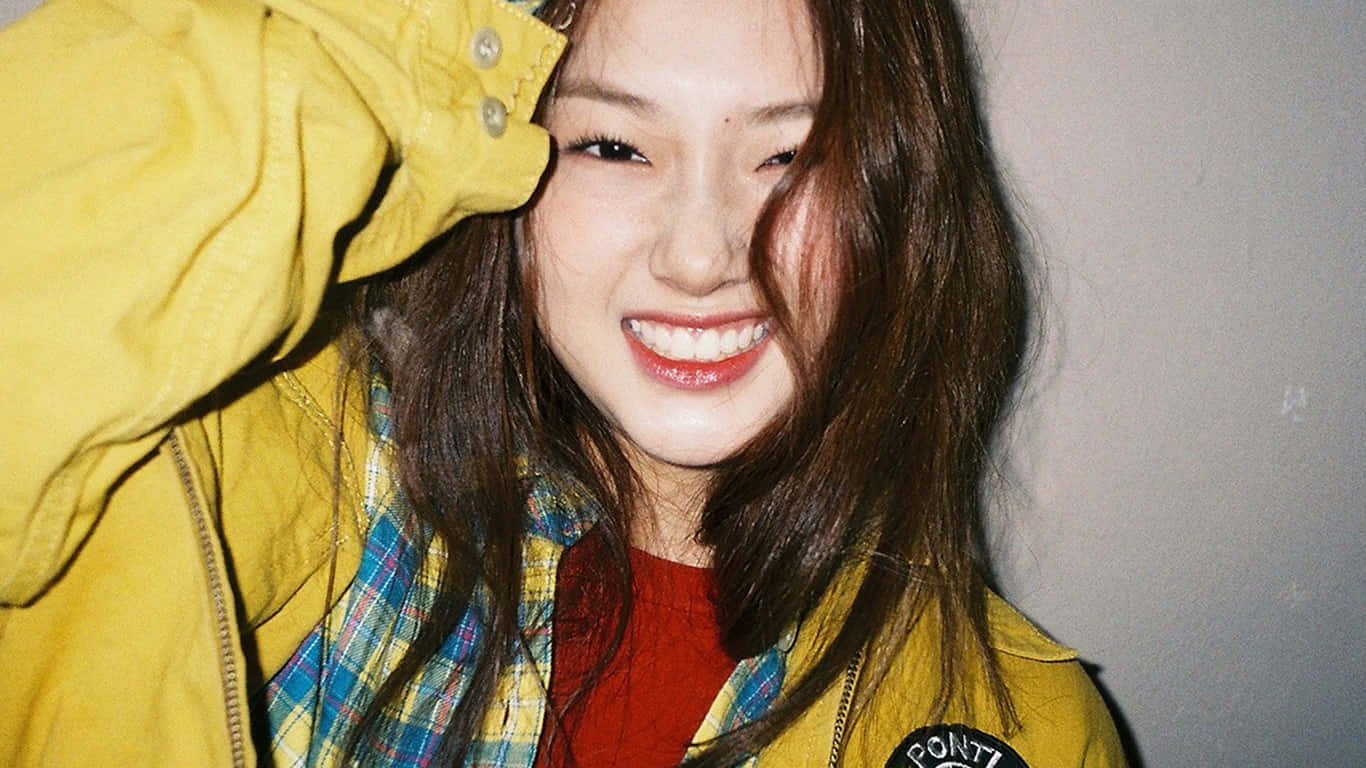  壁紙 — 一位長髮棕色的年輕女性，穿著黃色外套和格紋襯衫，在素色牆前燦爛微笑。