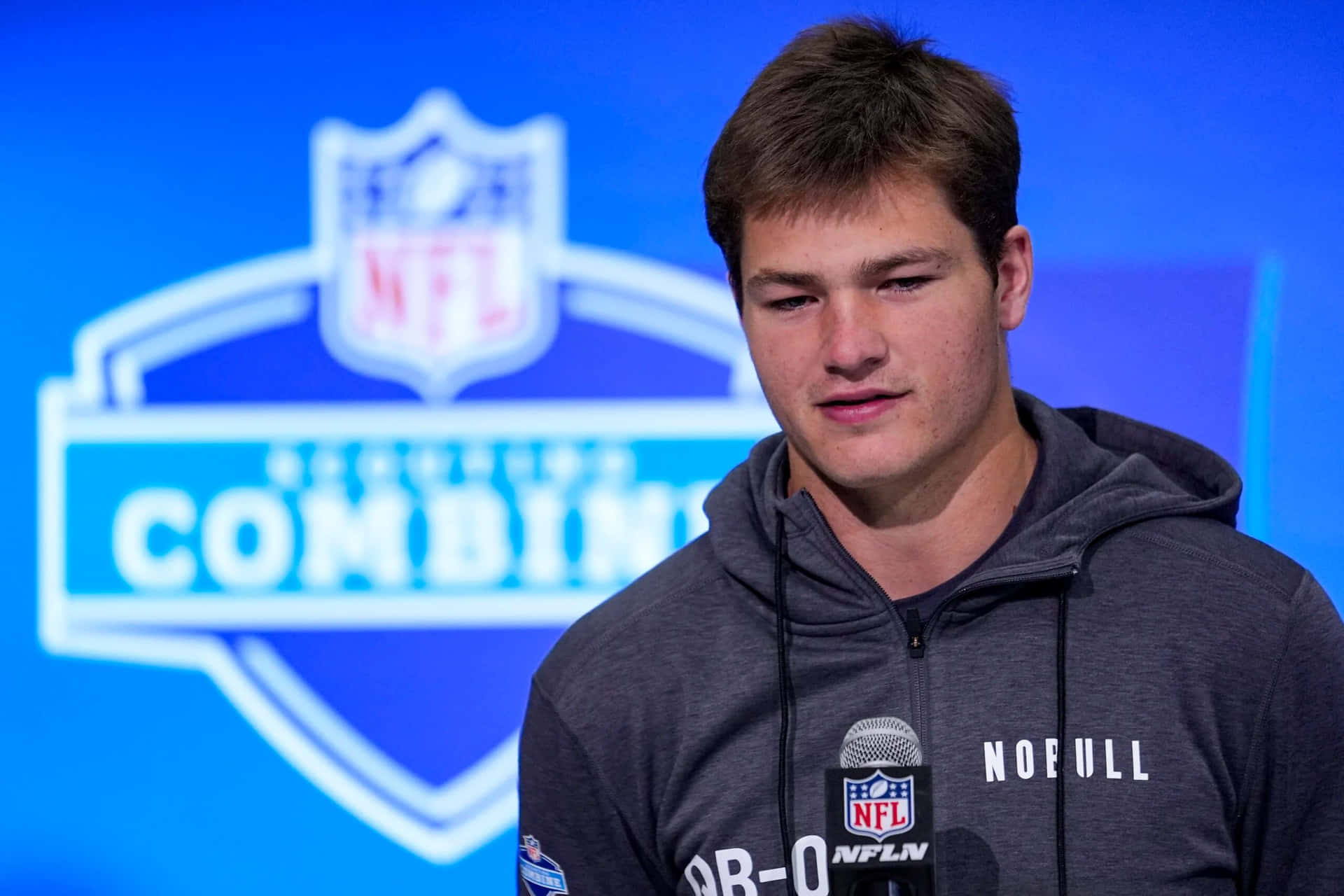  壁紙 — 年輕美式足球員身穿灰色連帽上衣，在NFL選秀聯合試訓記者會上發言，背景有標誌。