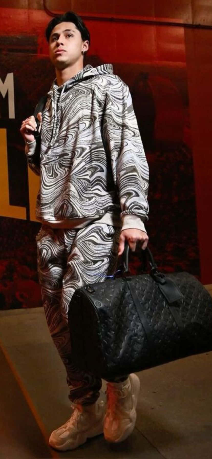  壁紙 — 一名身穿黑白漩渦圖案連帽上衣和長褲的年輕男子，在溫暖燈光下室內站立，攜帶一個黑色絎縫行李袋。
