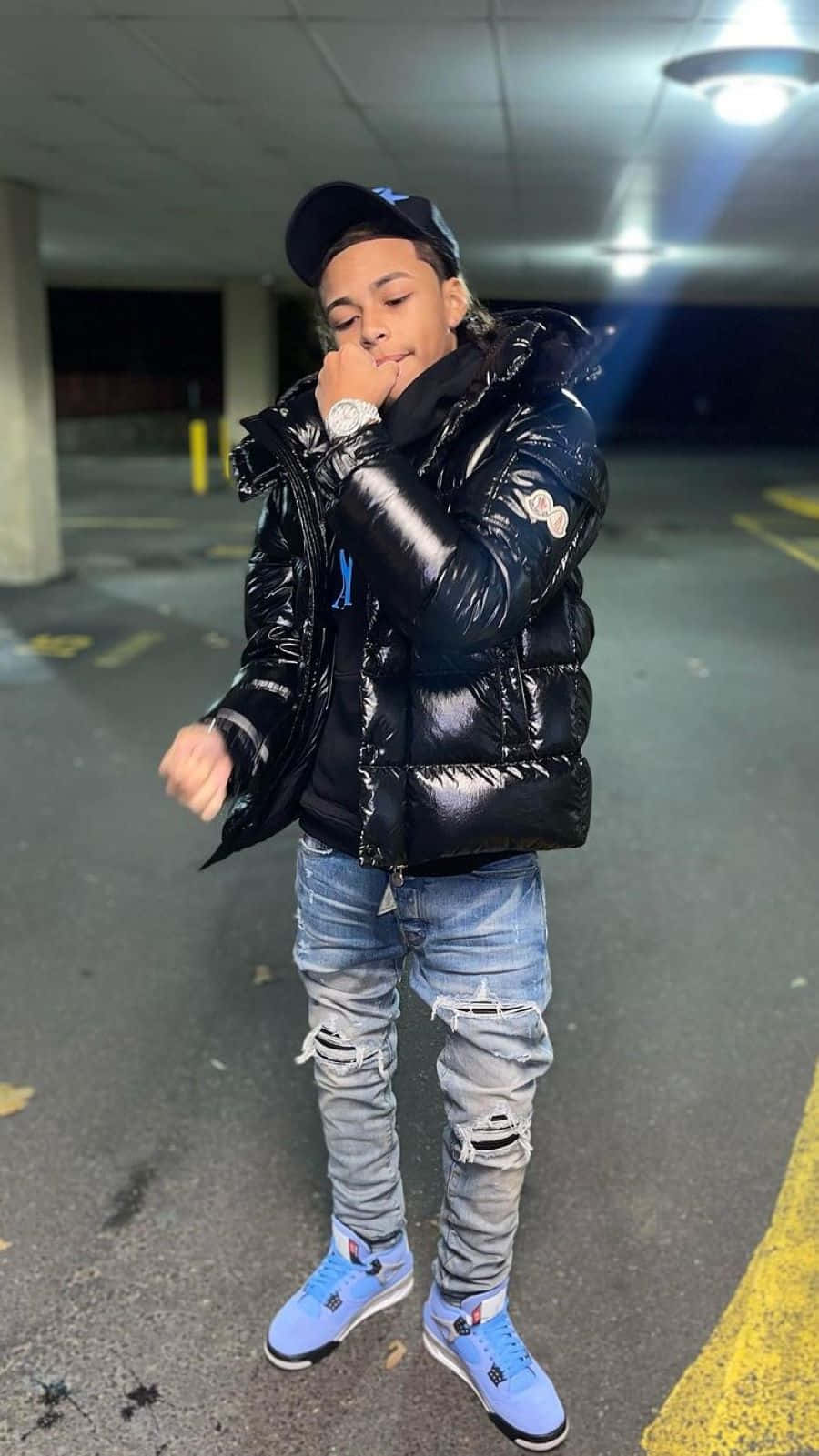  壁紙 — 一名年輕男子身穿閃亮黑色羽絨外套和破洞牛仔褲，在夜間光線昏暗的停車場自信地擺姿勢。