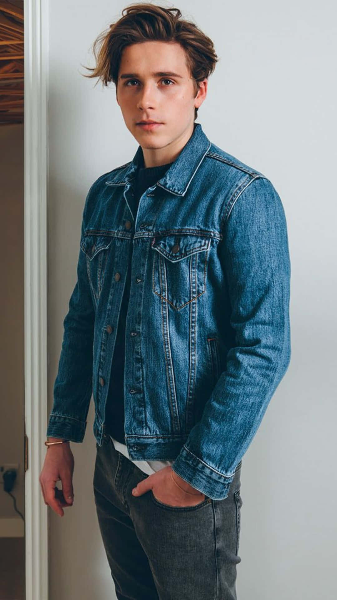  壁紙 — 一位年輕男子，棕色亂髮，身穿經典藍色牛仔外套，內搭黑色襯衫和深色牛仔褲，在室內靠著白色牆壁。