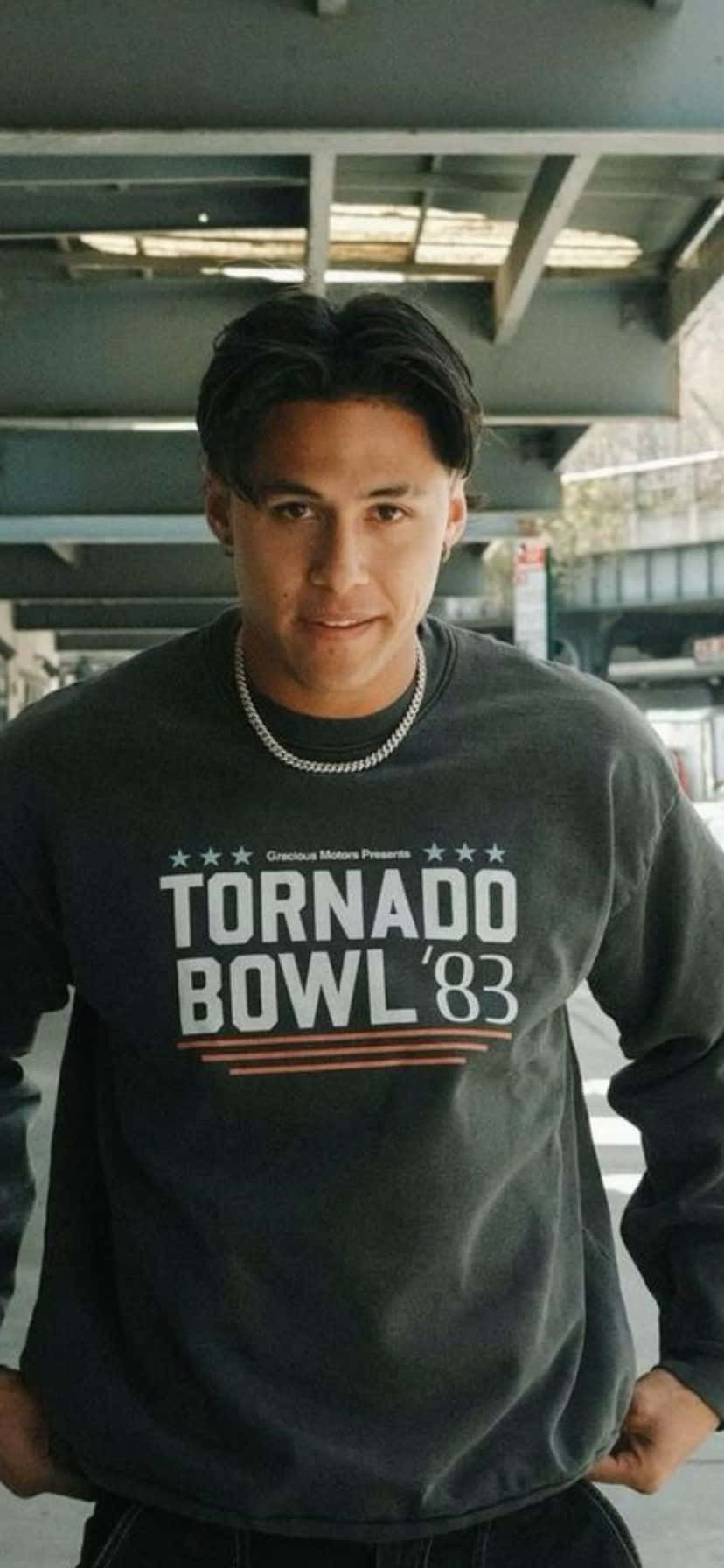  壁紙 — 一名身穿深色連帽上衣、印有 'TORNADO BOWL '83' 字樣的年輕男子自信地站在城市天橋下。