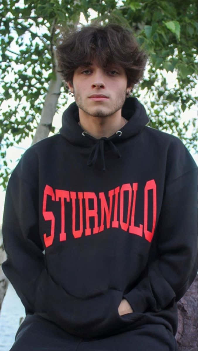  壁紙 — 一個年輕男子，一頭凌亂的黑髮，穿著一件黑色連帽衫，上面有粗體的紅色「STURMILO」文字，坐在戶外枝葉茂密的樹下。