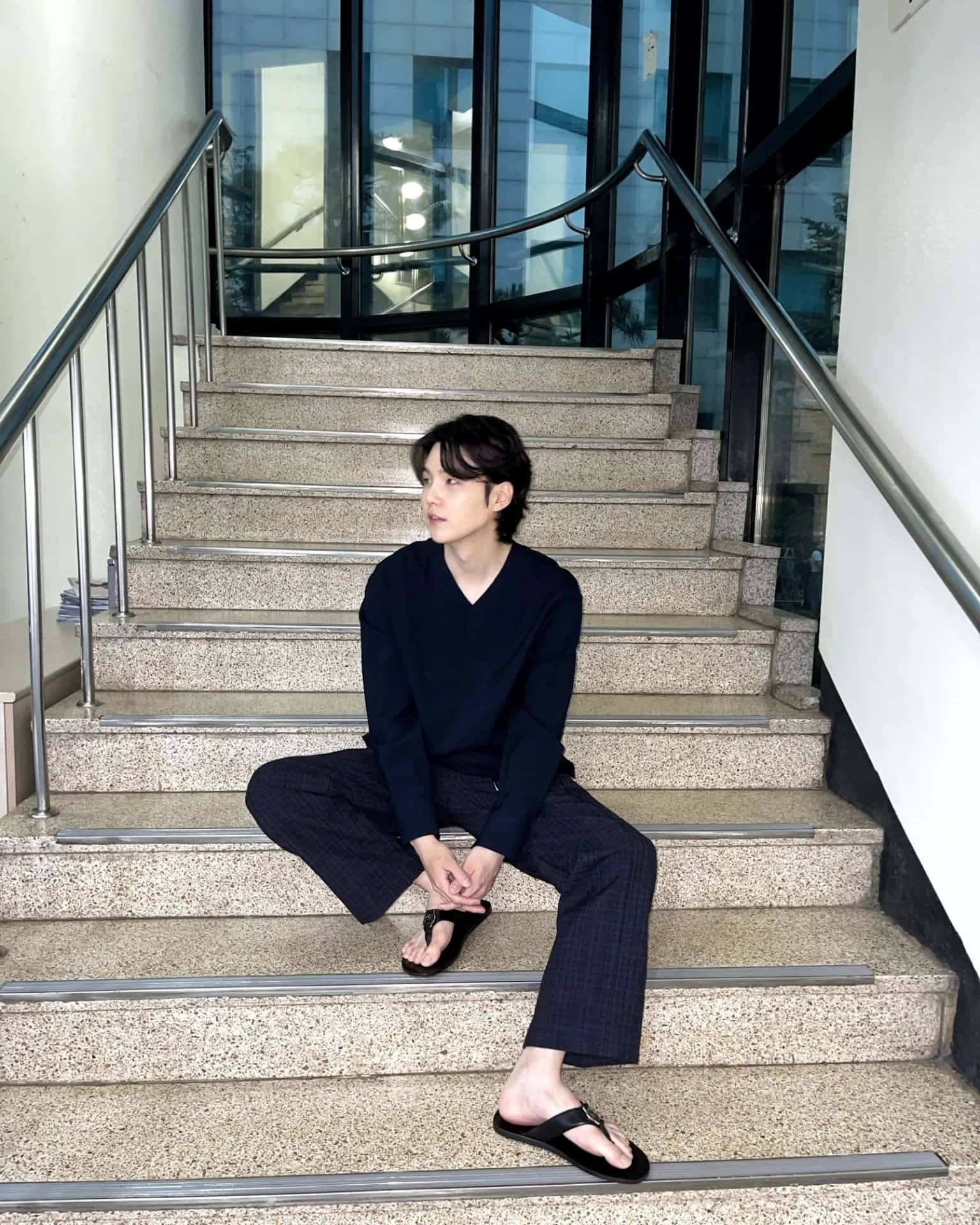  壁紙 — 一名身穿深色衣服的年輕男子，沉思地坐在現代石階上，階梯配有玻璃牆和金屬欄杆。