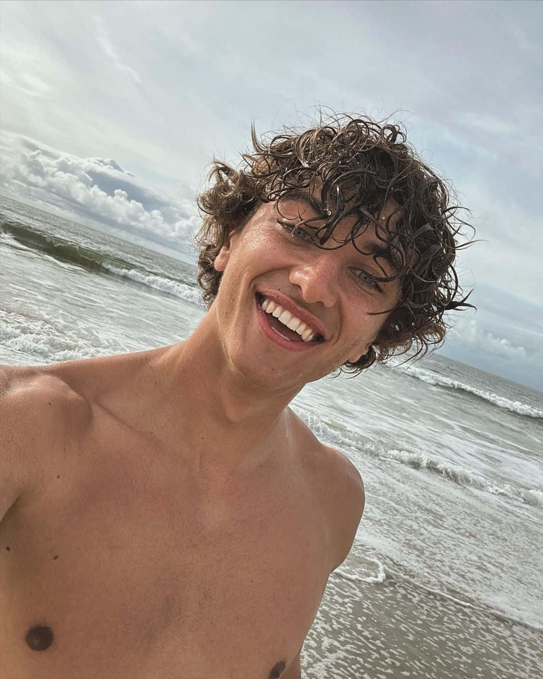  壁紙 — 一個年輕男子濕漉漉的捲髮，在陰天的海灘上，身後海浪翻滾，對著鏡頭開懷大笑。