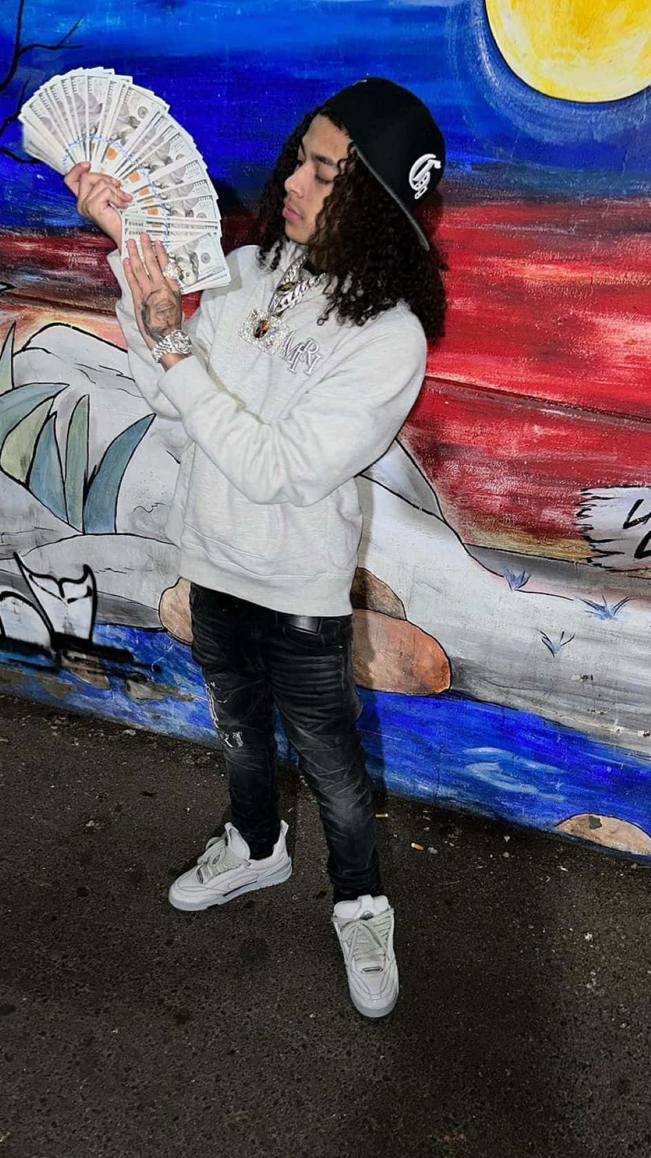  壁紙 — 一個捲髮年輕男子手持一疊現金，站在色彩繽紛的塗鴉壁畫前，背景是明亮的黃色太陽。