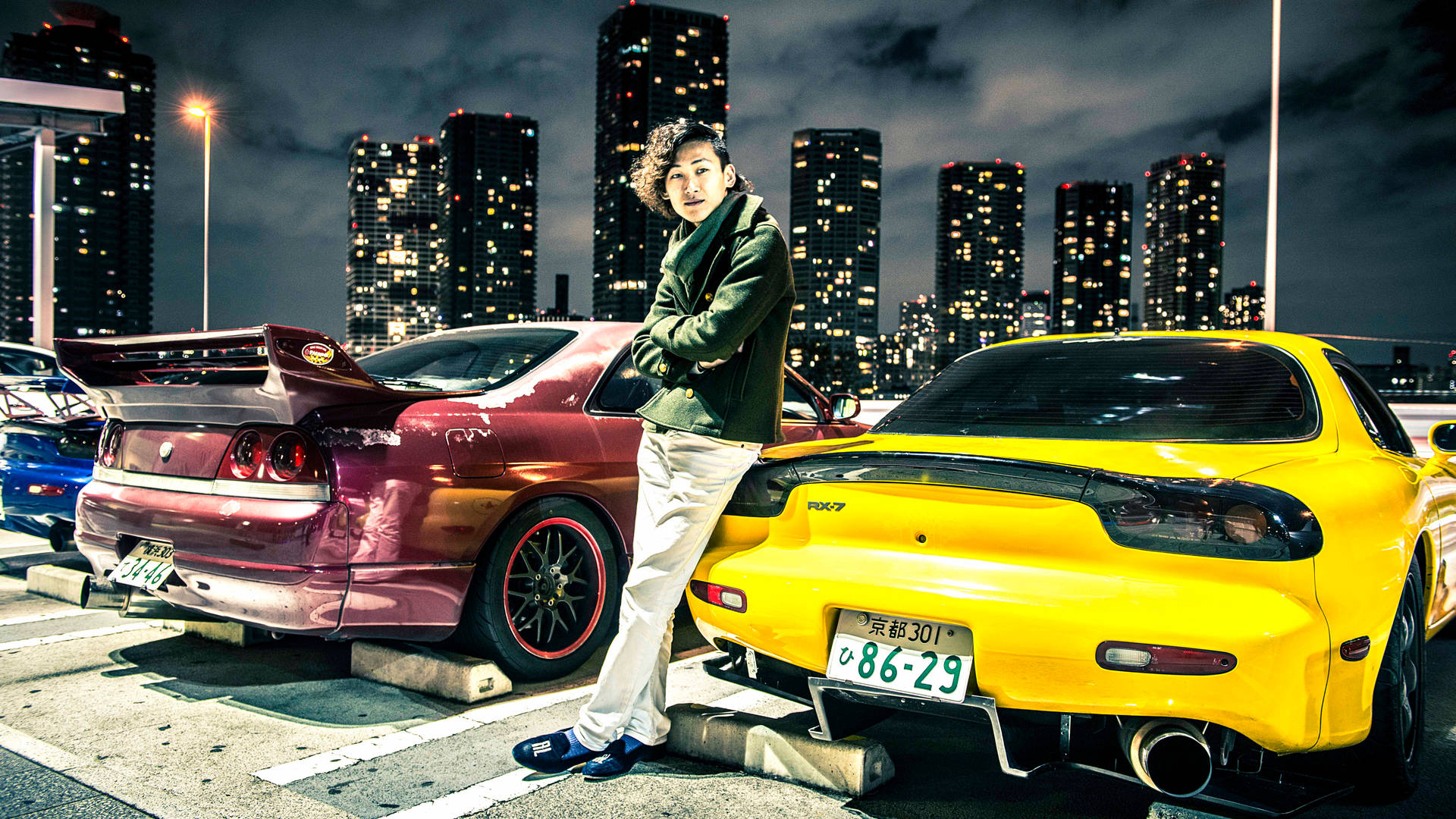 年輕男子在夜晚的城市停車場中靠在兩輛跑車之間。 — JDM汽車 壁紙