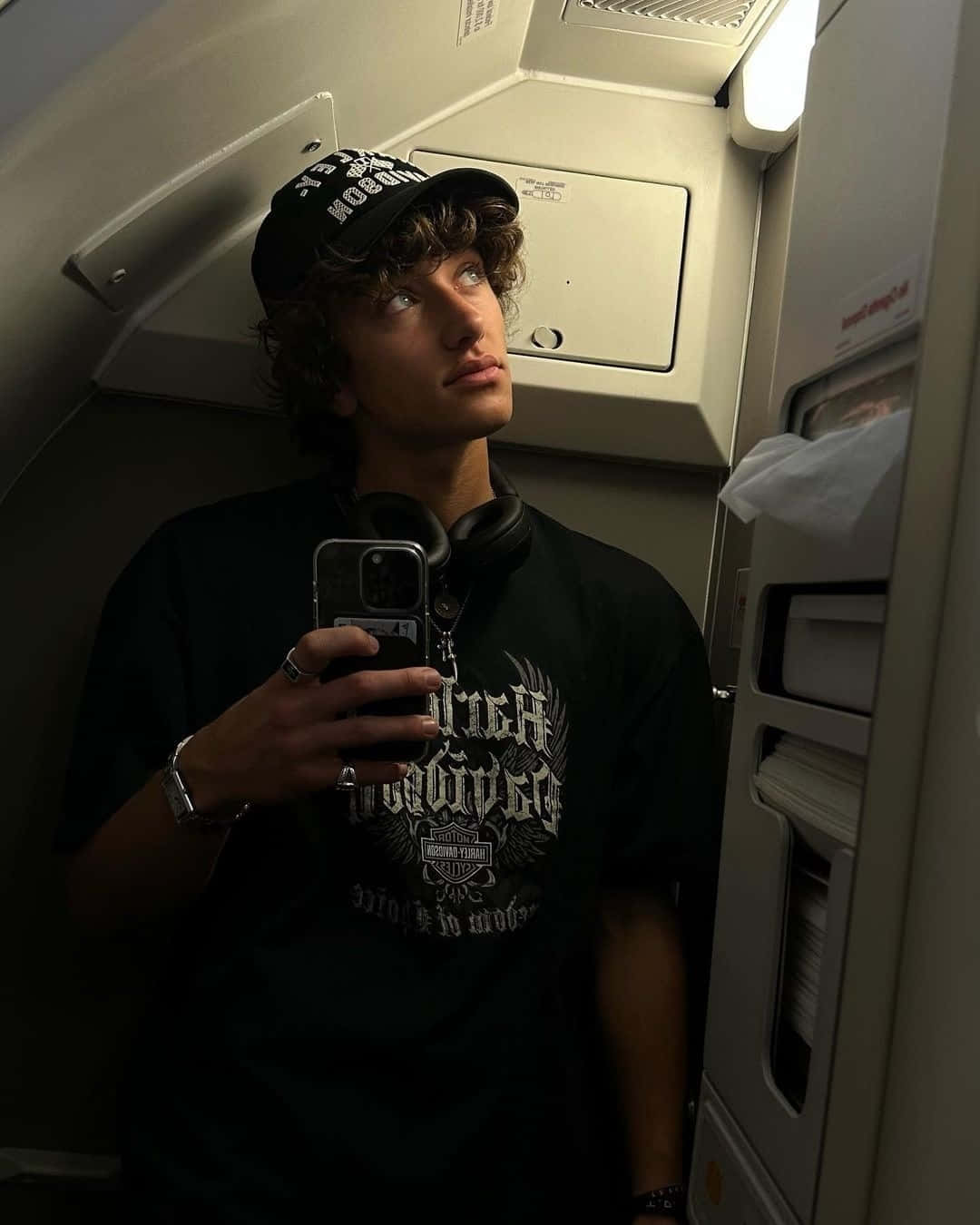  壁紙 — 一位捲髮年輕男子在飛機艙內戴著黑色帽子和耳機對著鏡子自拍。