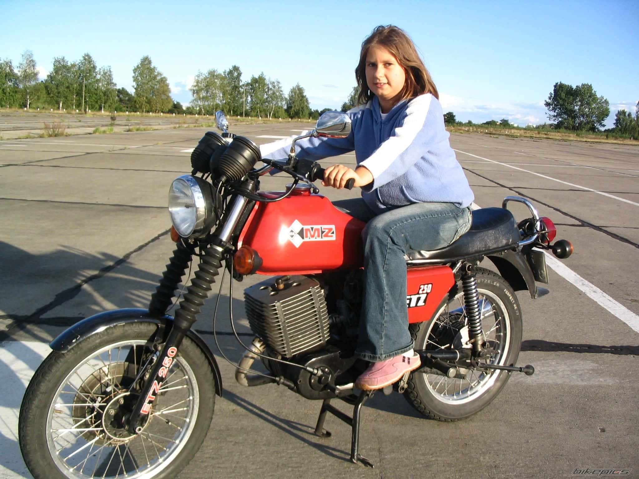 Mz Motorcycle 桌布 壁紙 — 一個年輕女孩自信地坐在一輛紅色復古機車上，背景是陽光普照的停車場和樹木。