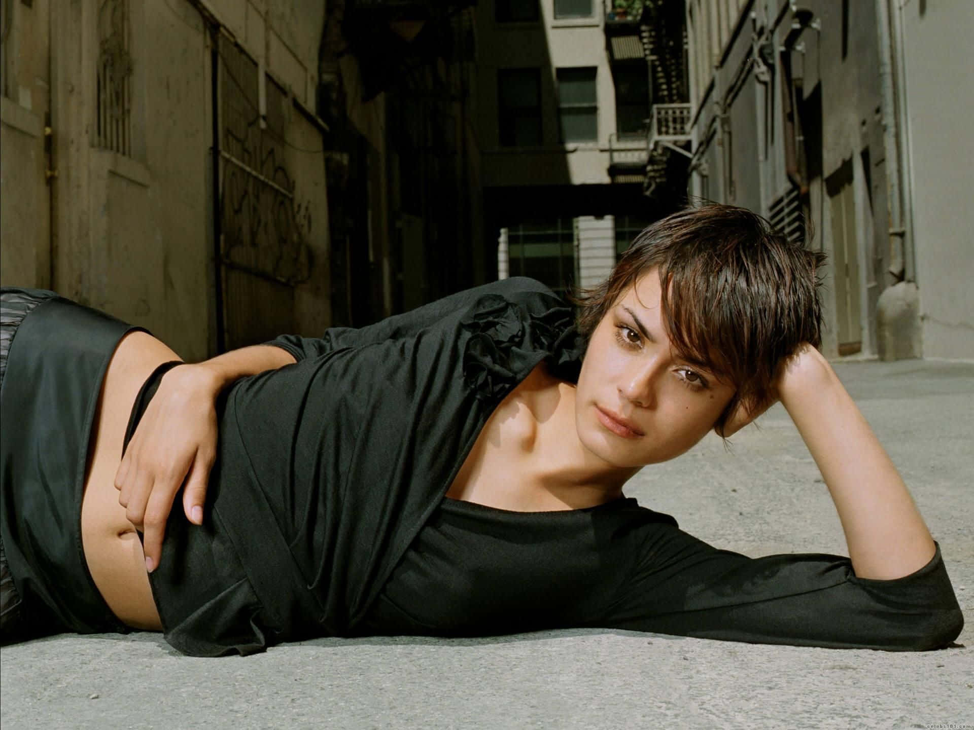 Shannyn Sossamon 桌布 壁紙 — 一位短黑髮年輕女性躺在陽光普照的城市小巷地上，直視鏡頭，表情若有所思。
