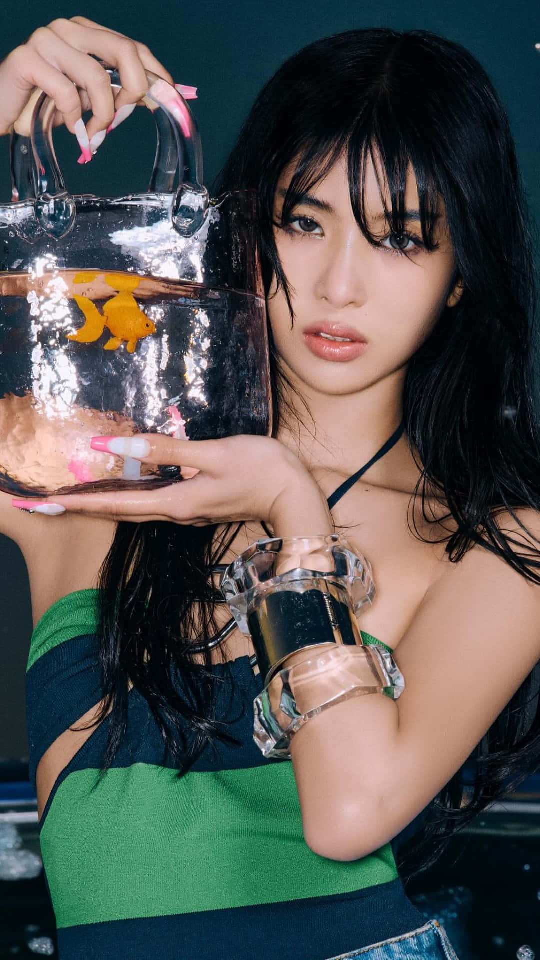  壁紙 — 一位深色頭髮的年輕女性手持一個透明手提包，內有金魚和水，佩戴大膽的飾品和綠色上衣。