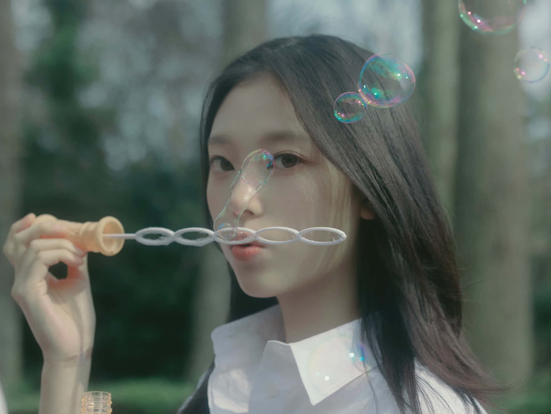  壁紙 — 一位年輕女性在陽光明媚的森林中吹出彩虹色的泡泡，背景是柔焦的樹木。
