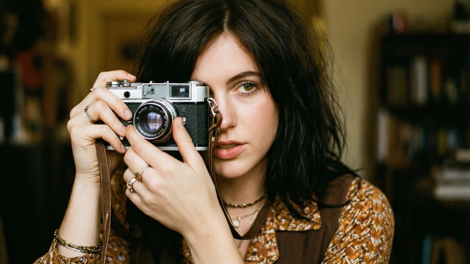 Vintage Photographer 壁紙 — 一位深色頭髮的年輕女性在一個光線溫暖、有書架的房間裡，將一部復古銀色相機舉到眼前。