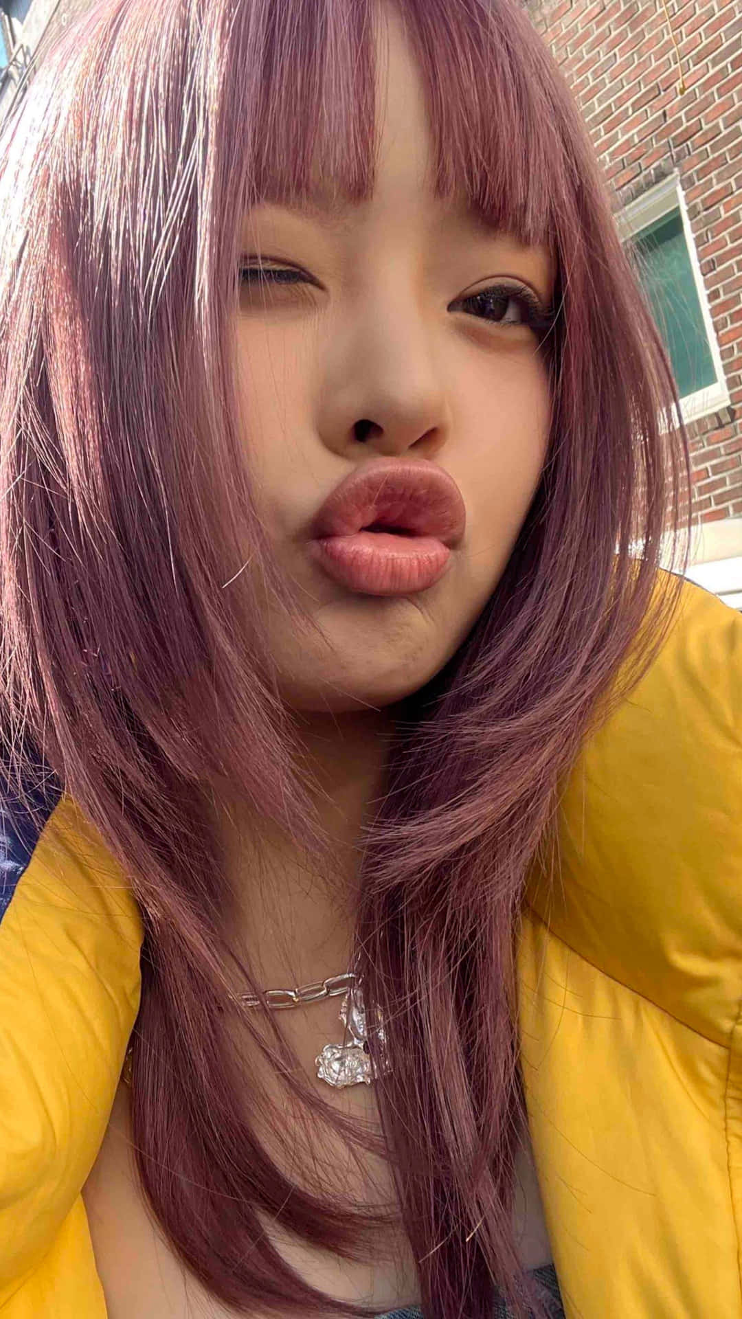  壁紙 — 一位年輕女性擁有充滿活力的紫色頭髮和黃色外套，對著磚牆嘟嘴拍一張俏皮的自拍照。