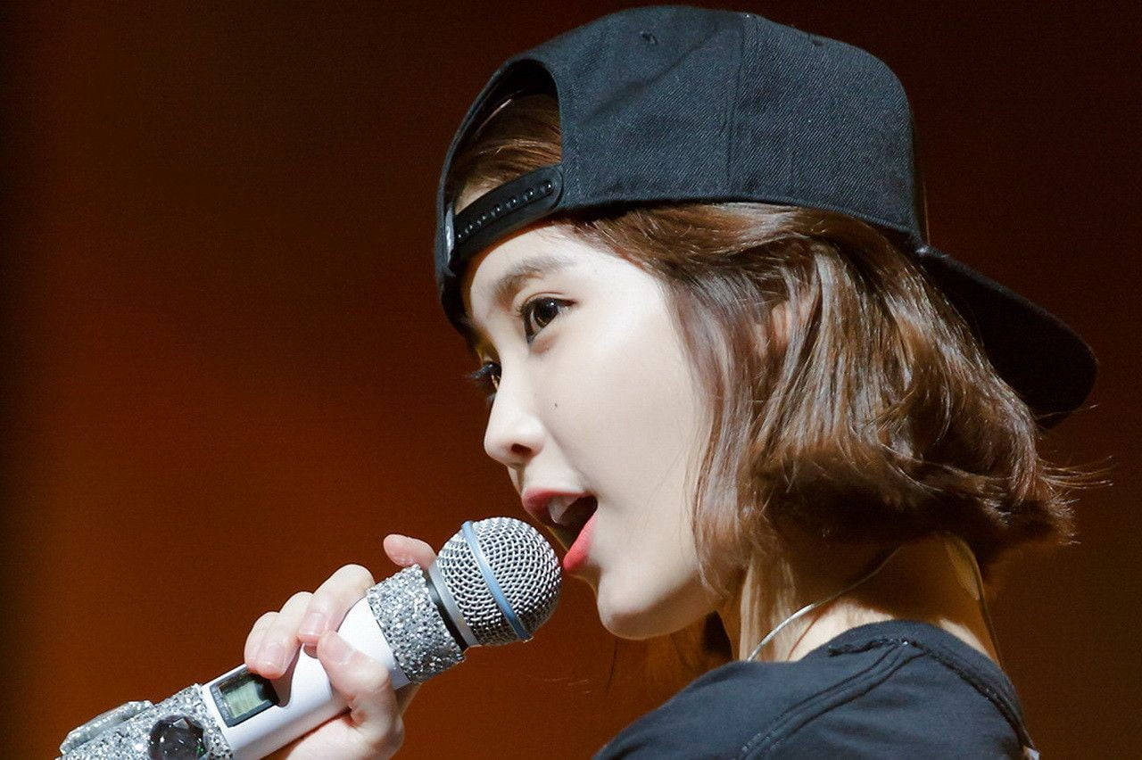 年輕女子在舞台上戴黑色帽子對著麥克風唱歌 — K Pop 壁紙