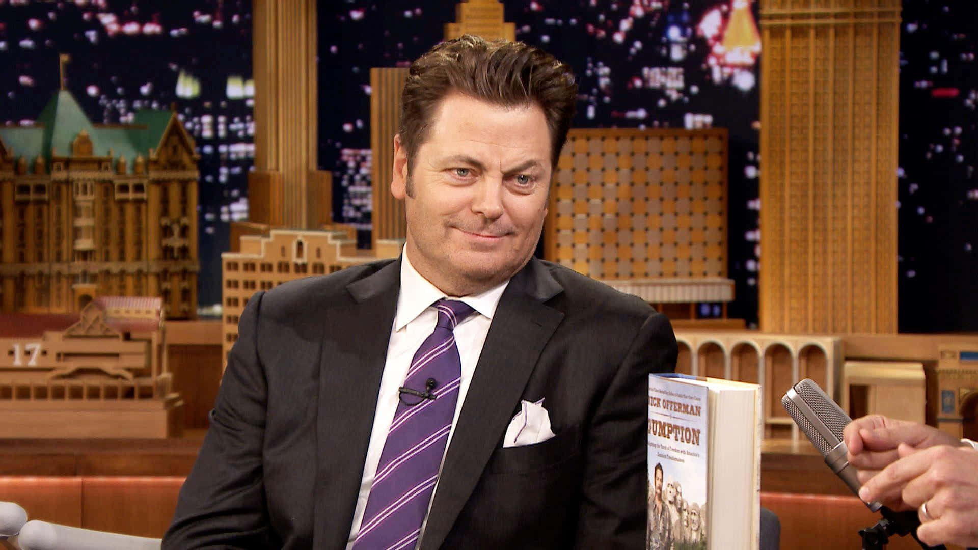 nick offerman 桌布 壁紙 — Nick Offerman 身穿俐落西裝，在設有微型城市背景的脫口秀舞台上微笑著手持自己的書。