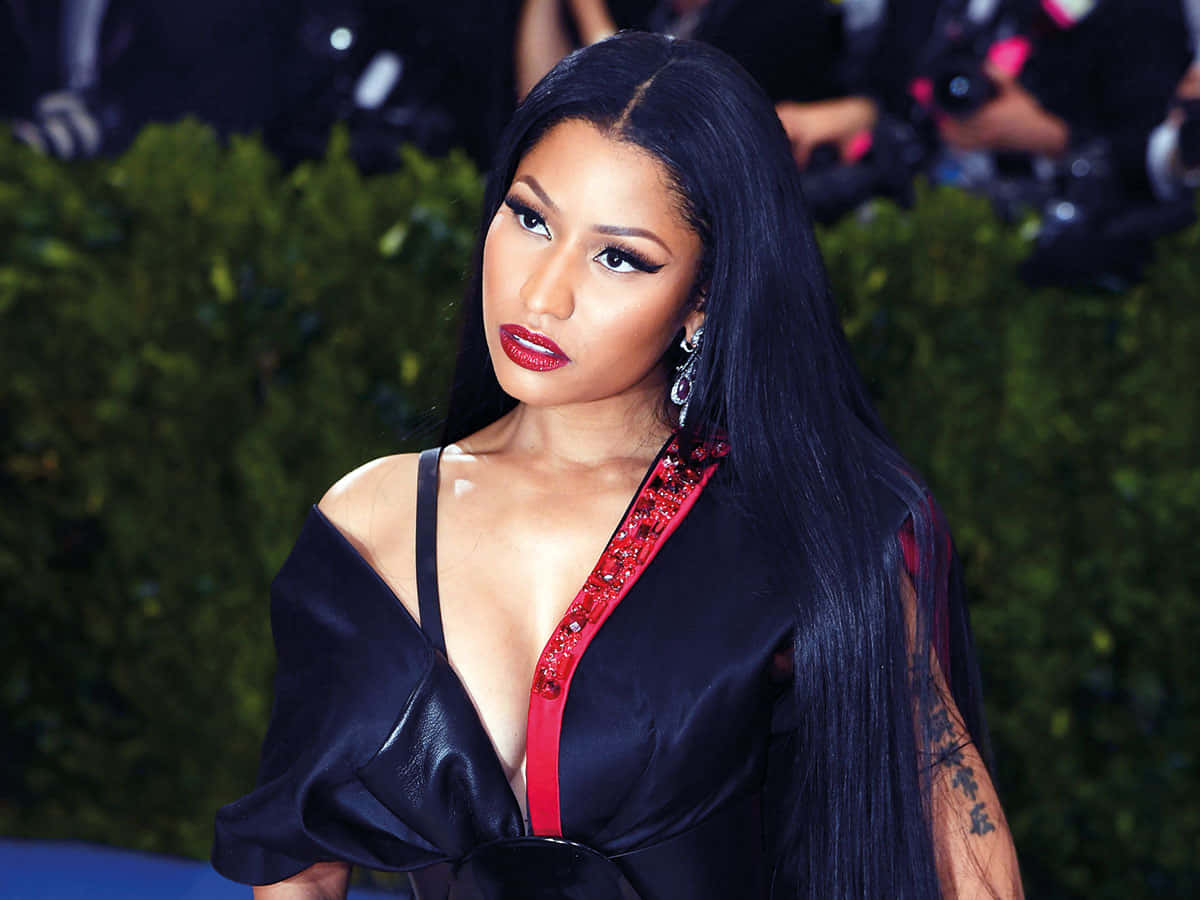 妮琪·米娜 桌布 壁紙 — Nicki Minaj 自信地擺姿勢，身穿黑色緞面連衣裙，配有紅色裝飾肩帶，背景是綠色樹籬。