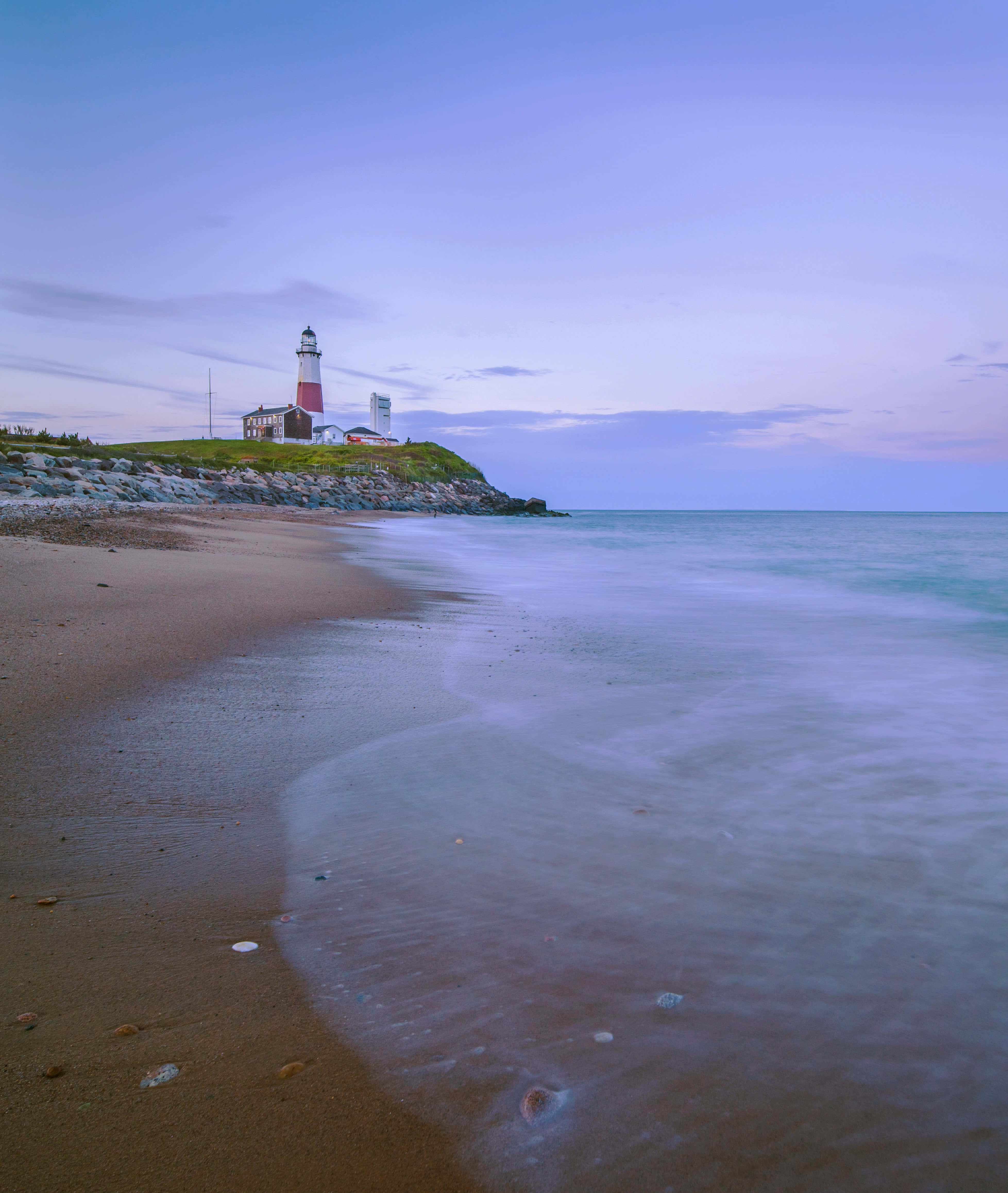 Coastal Granddaughter 桌布 壁紙 — 寧靜的黃昏海灘場景，溫柔的海浪拍打著海岸，一座紅白相間的燈塔矗立在長滿青草的懸崖上。