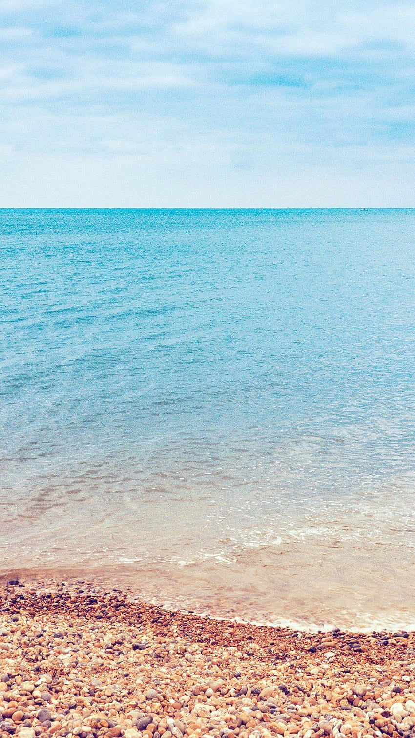  壁紙 — 寧靜的卵石海灘在柔和藍天與薄雲下，與平靜的碧綠海水相遇。