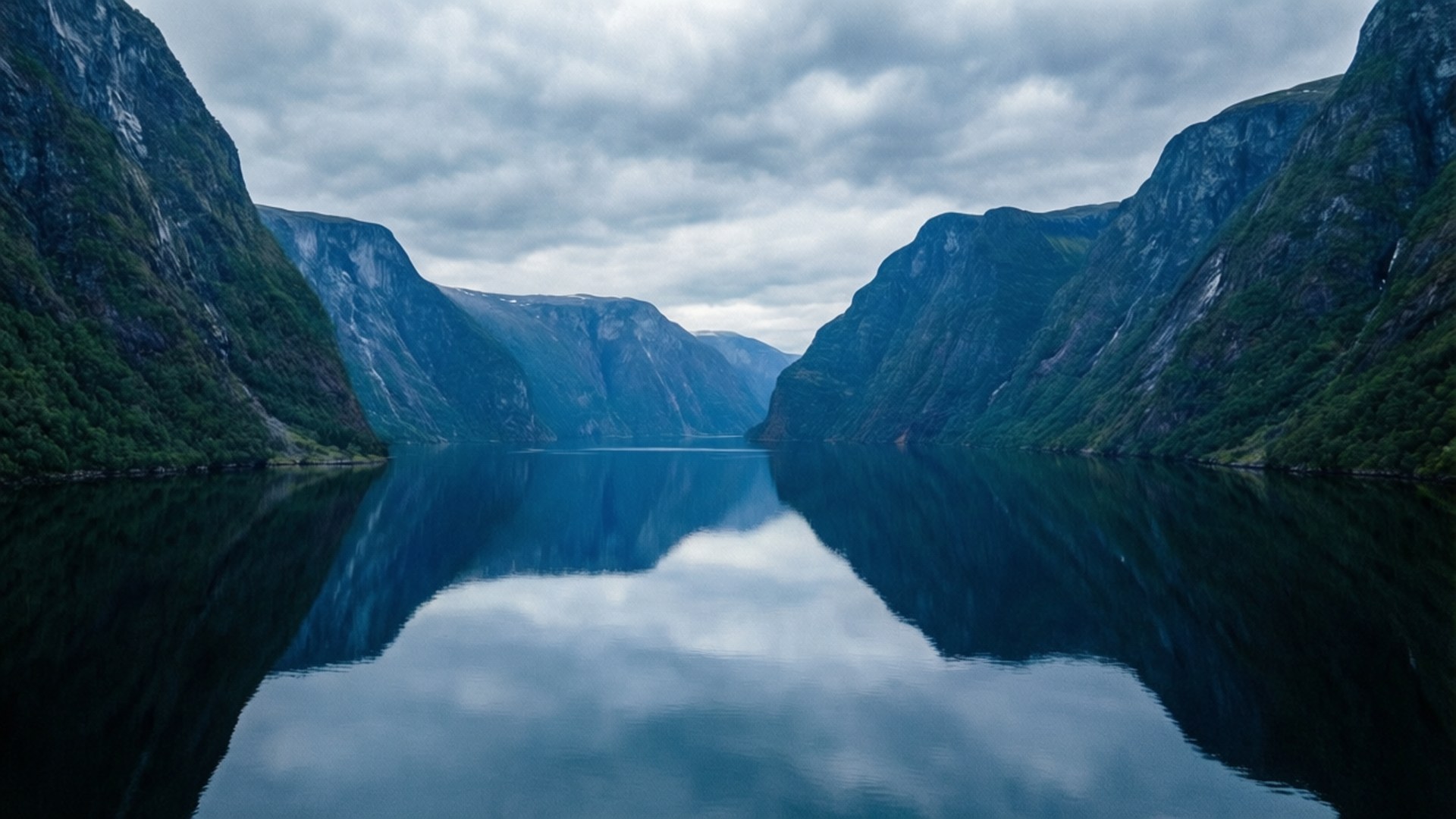 Moody Fjord Reflection 壁紙 — 寧靜的峽灣在如鏡的水面上倒映著高聳的綠色懸崖和多雲的天空。