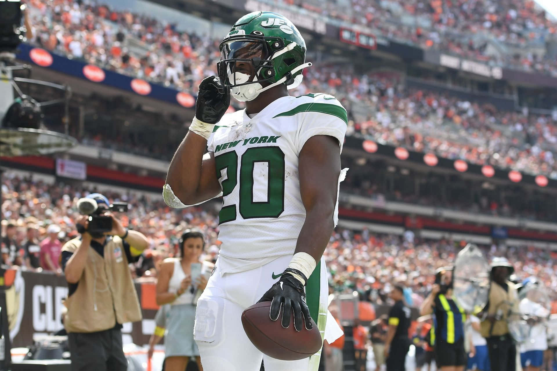  壁紙 — 一位身穿白色和綠色球衣的紐約噴射機美式足球員，在陽光明媚的球場上抱著足球，高舉手臂慶祝勝利。