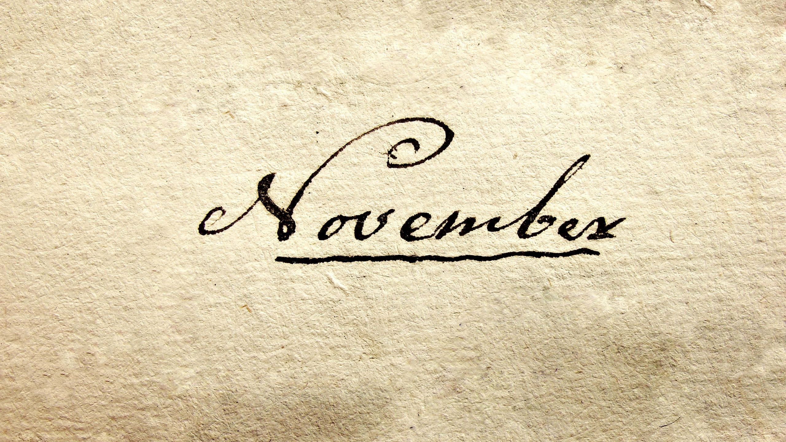 November Desktop 桌布 壁紙 — 「November」這個詞以黑色墨水優雅地書寫在帶有細微瑕疵的紋理米色紙上。