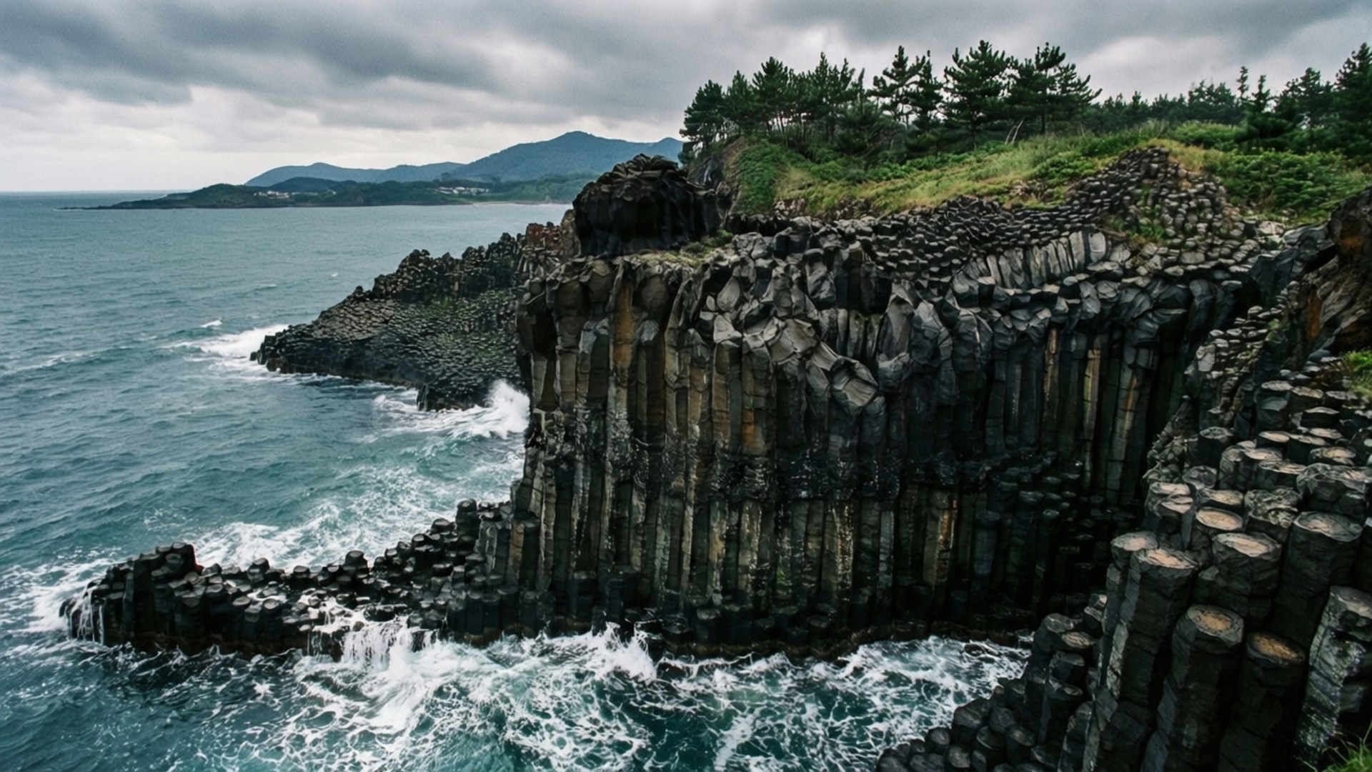 Jusangjeolli Cliff Columns 壁紙 — 海浪拍打高聳的深色玄武岩柱，在多雲的天空下形成崎嶇的海岸懸崖。