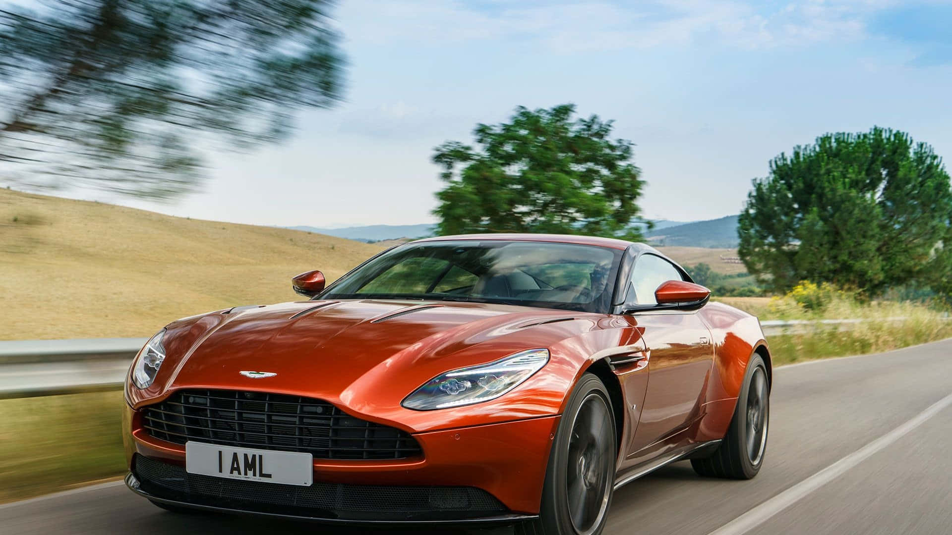 Aston Martin Db11 桌布 壁紙 — 一輛流線型橙色 Aston Martin 在蜿蜒的鄉村道路上疾駛，背景是模糊的綠樹和金色山丘，上方是明亮的藍天。