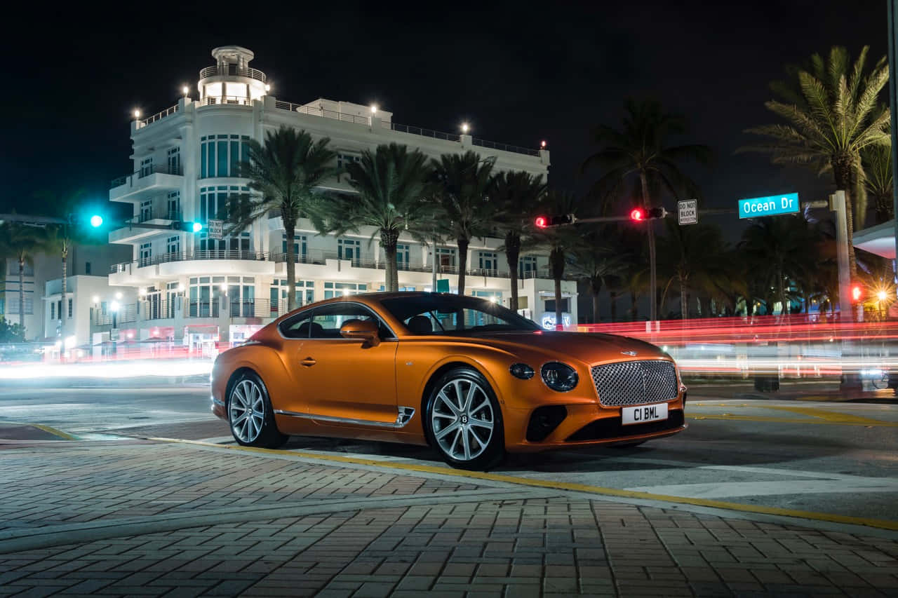Bentley Continental Gt 桌布 壁紙 — 一輛閃亮的橙色賓利 Continental GT 在夜間停泊在海洋大道上，棕櫚樹和模糊的城市燈光掠過。