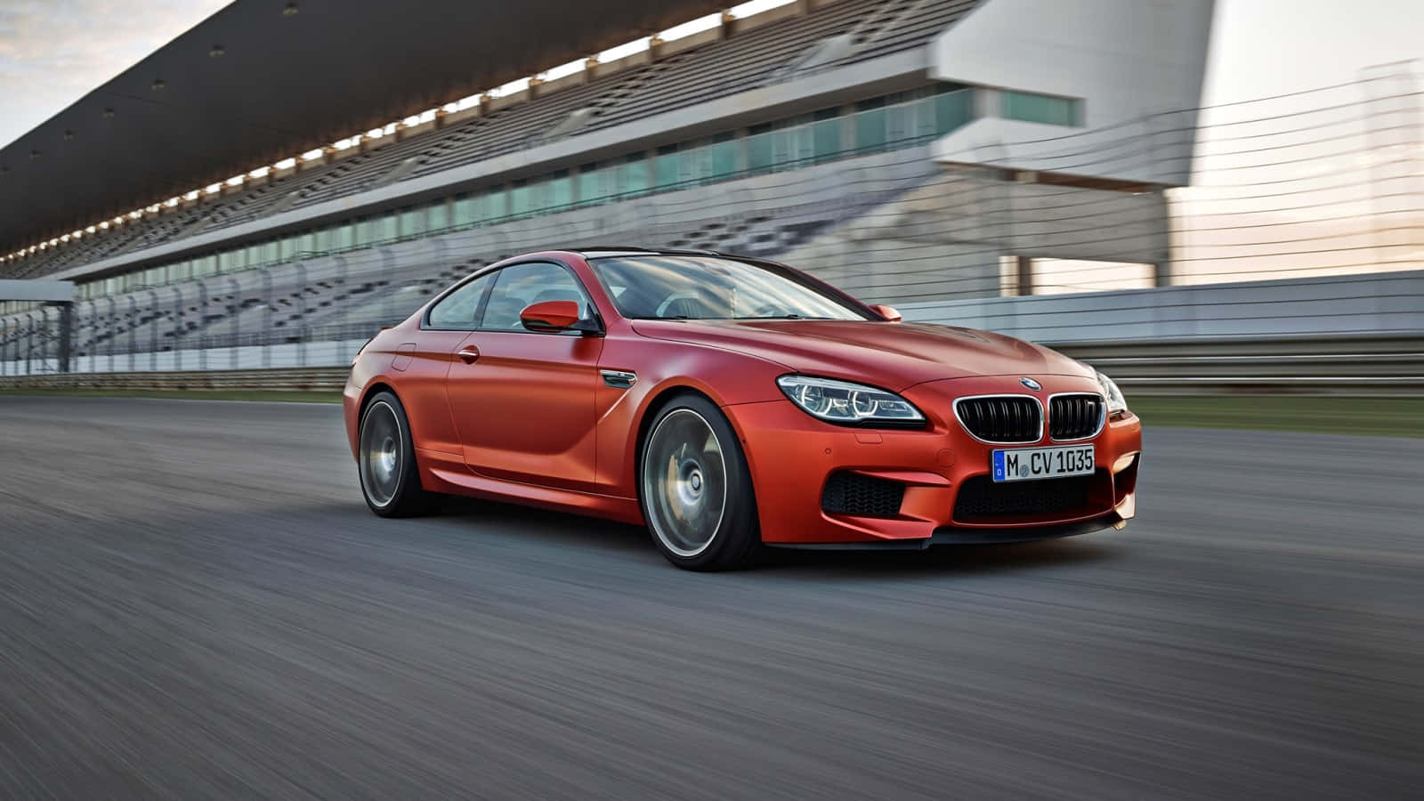 Bmw M6 桌布 壁紙 — 一輛充滿活力的橙色BMW M6雙門跑車在現代看台下高速駛過賽道，帶有動態模糊效果。