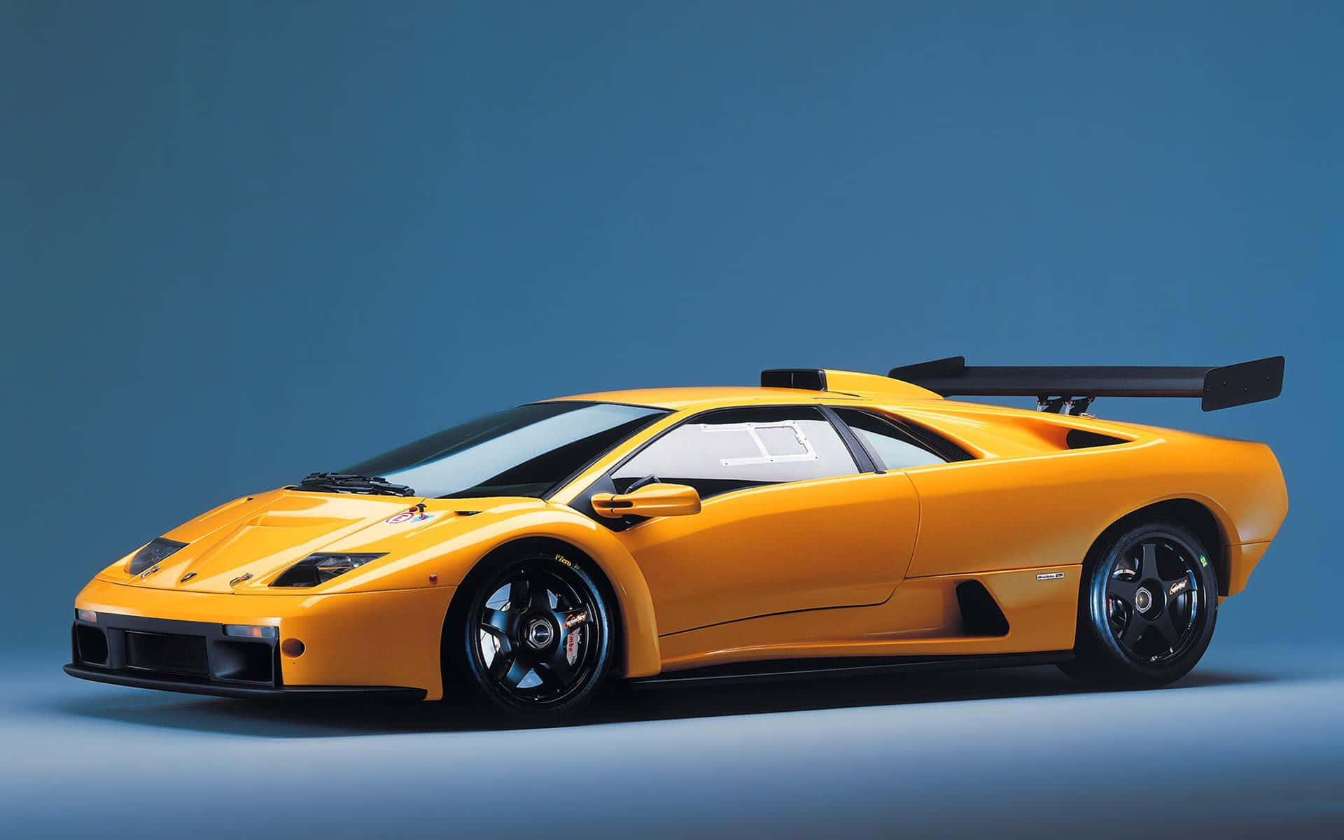 Lamborghini Diablo 桌布 壁紙 — 一輛充滿活力的橙色藍寶堅尼 Diablo SV 跑車，帶有黑色飾邊和大型後尾翼，在純藍色工作室背景前展示。