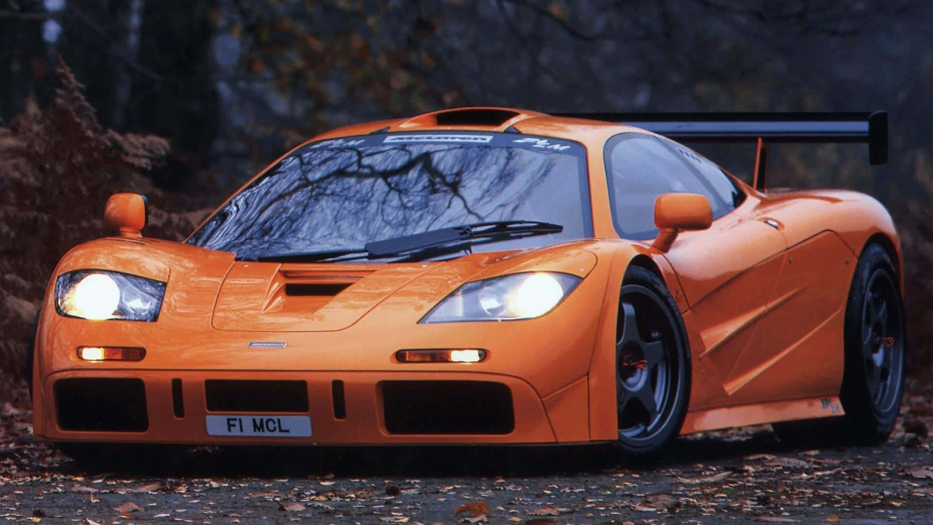Mclaren F1 桌布 壁紙 — 一輛充滿活力的橙色McLaren F1跑車，車頭燈閃爍，停泊在黃昏時分鋪滿落葉的森林小徑上。