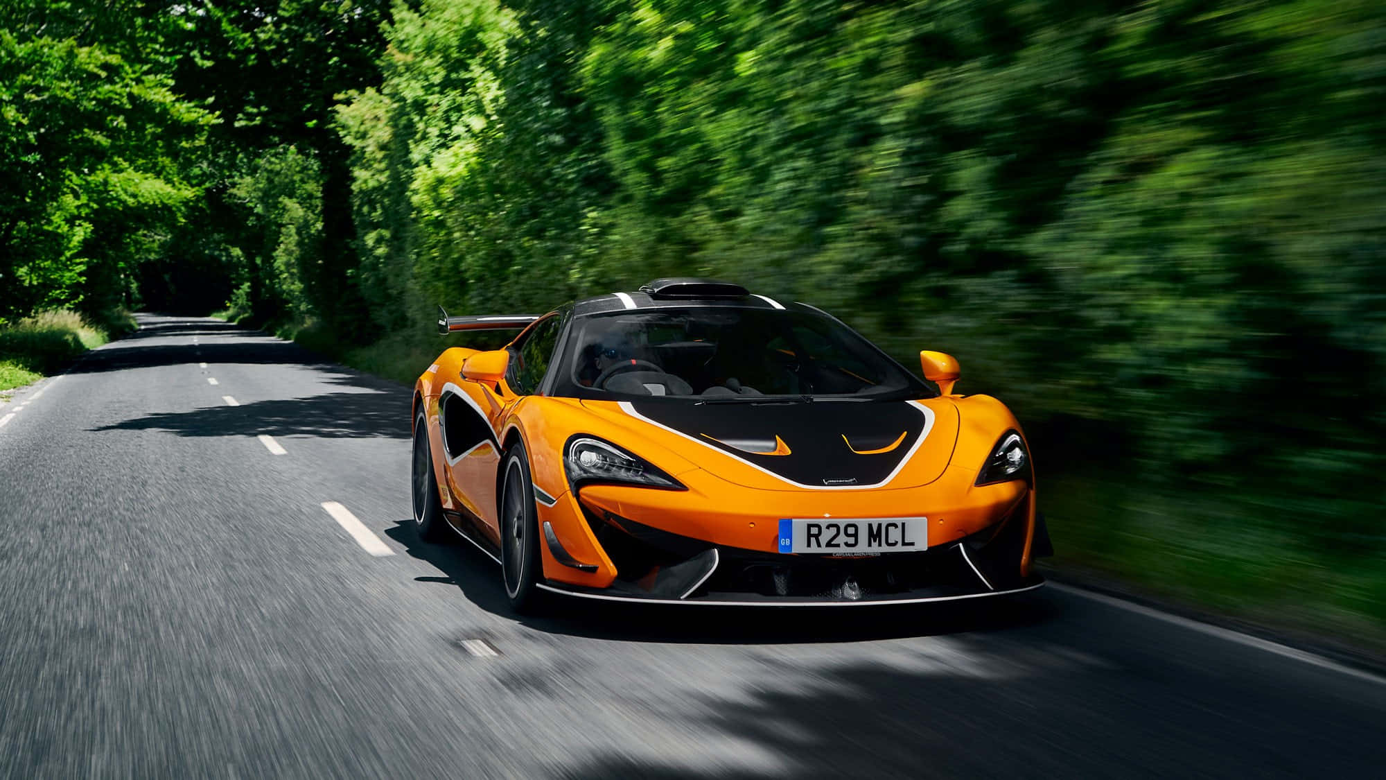 Mclaren 620r 桌布 壁紙 — 一輛鮮橙色的McLaren跑車在樹蔭大道上飛馳，動態模糊效果強調其速度。