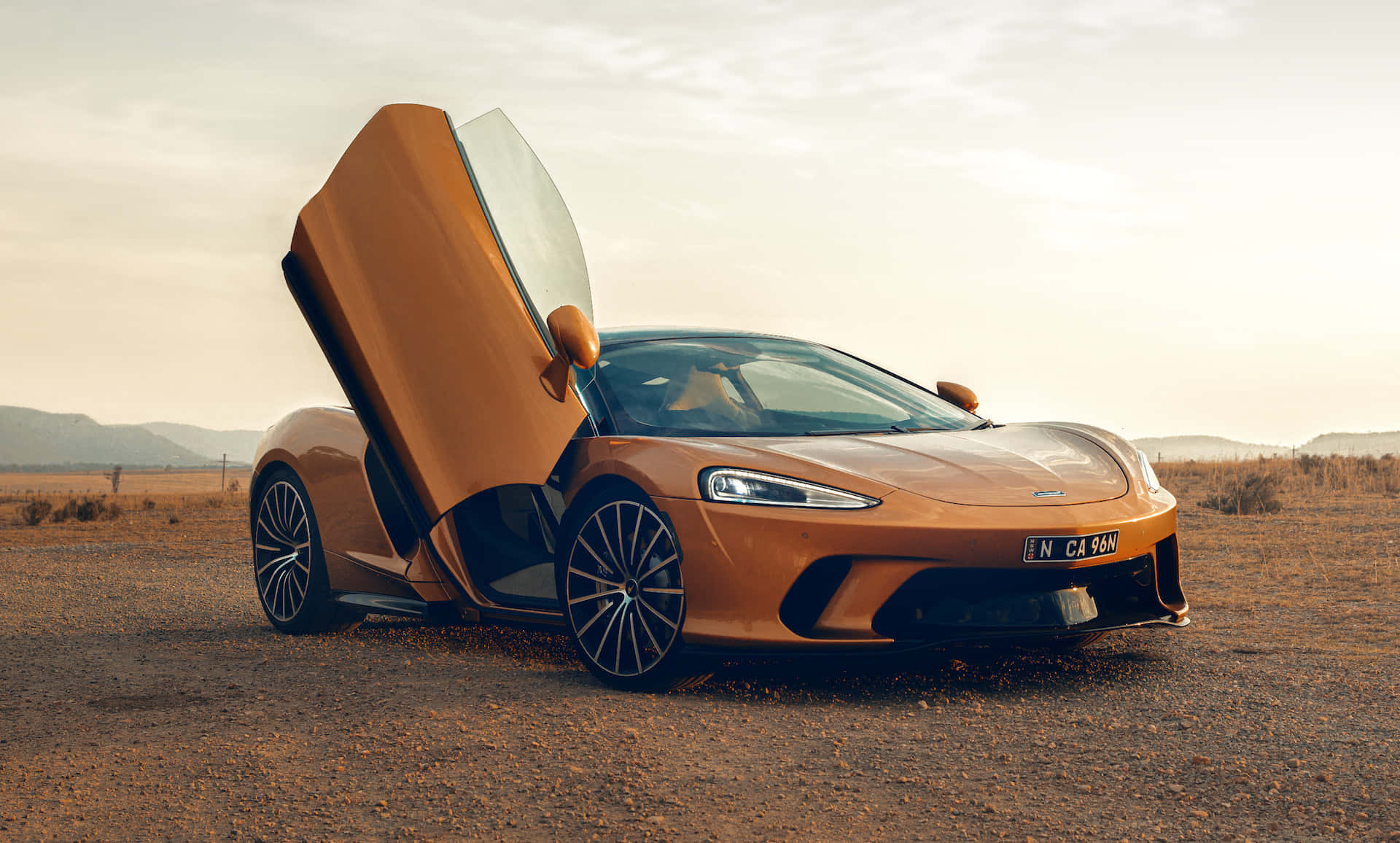 Mclaren Gt 桌布 壁紙 — 一台流線型的橙色McLaren跑車，蝴蝶門打開，停在塵土飛揚的沙漠公路上，天空呈現柔和的金色。