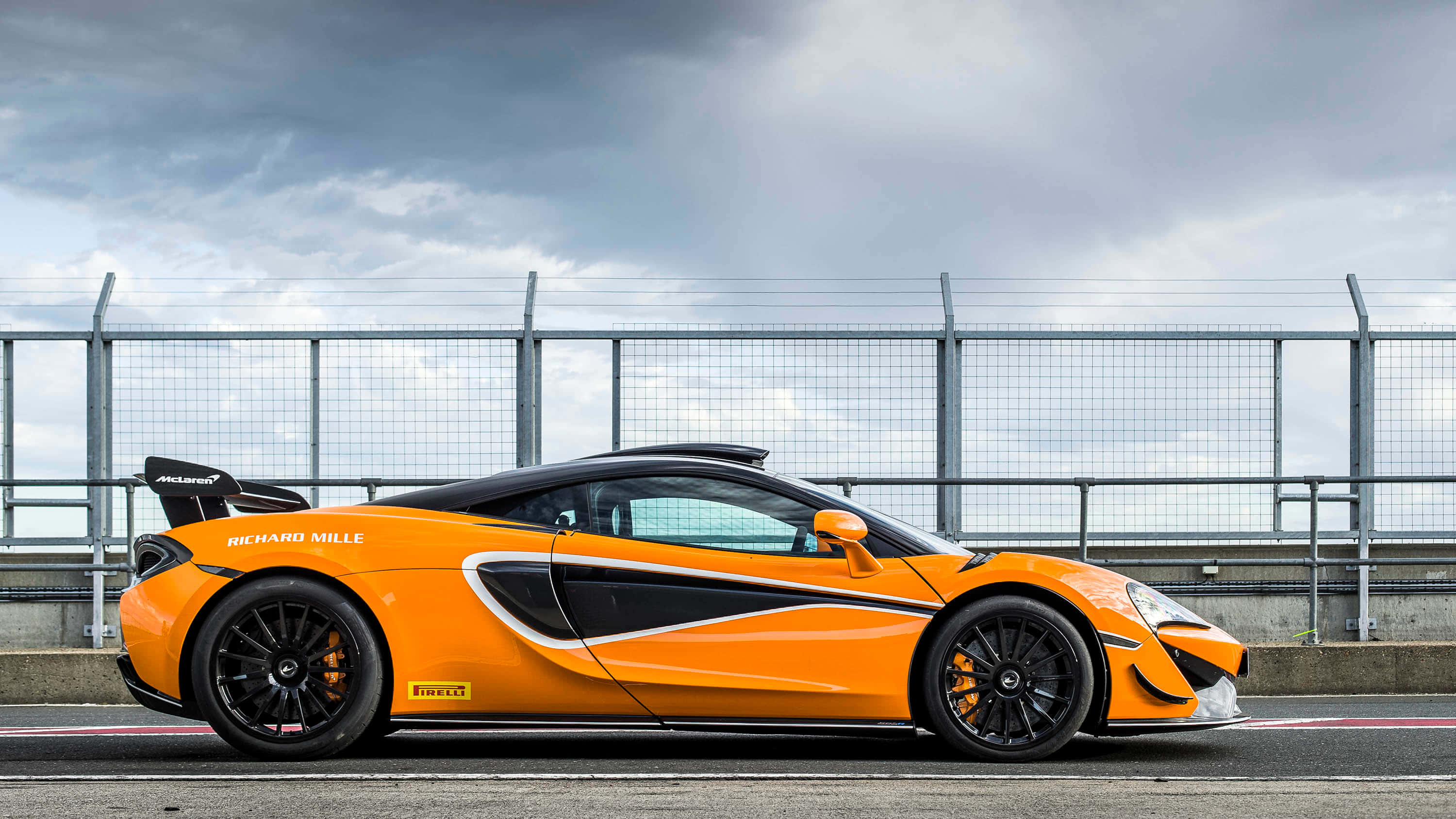 Mclaren 620r 桌布 壁紙 — 一輛充滿活力的橙色McLaren跑車，帶有黑色飾邊和賽車貼紙，在陰天下的賽道上蓄勢待發。