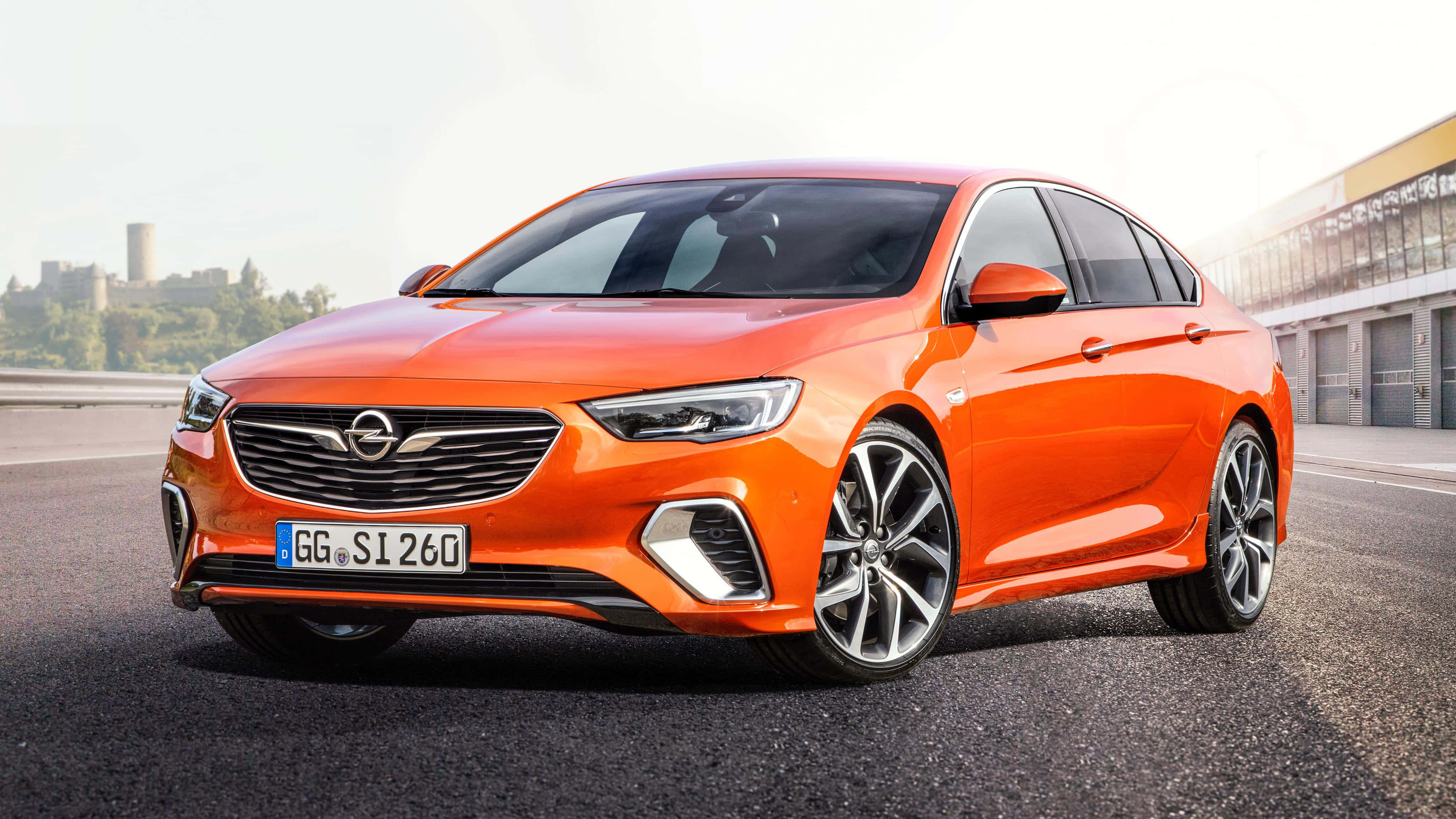 Opel Insignia 桌布 壁紙 — 一輛充滿活力的橙色Opel轎車在明亮的日光下，在瀝青路上閃閃發光，背景是一座城堡。