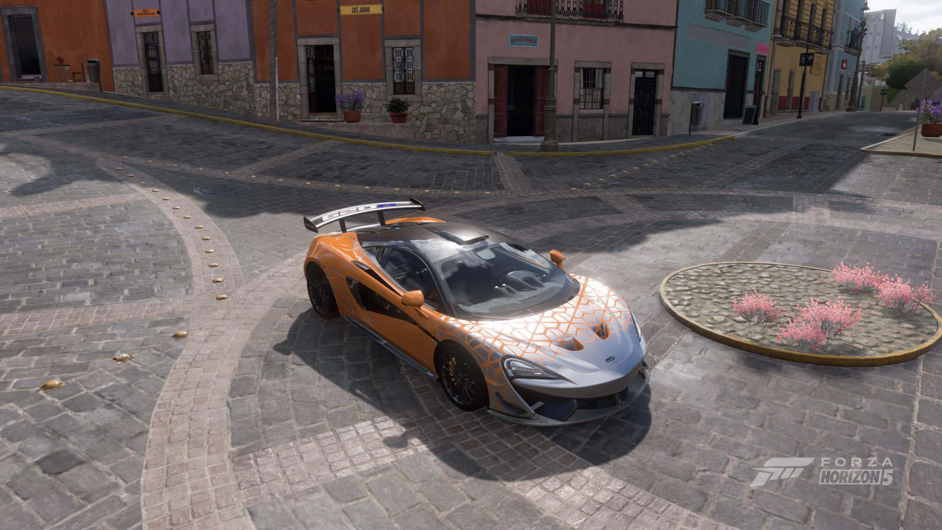 Mclaren 620r 桌布 壁紙 — 一輛充滿活力的橙色和銀色麥拉倫跑車停泊在鵝卵石廣場上，周圍是色彩繽紛的殖民時期建築。