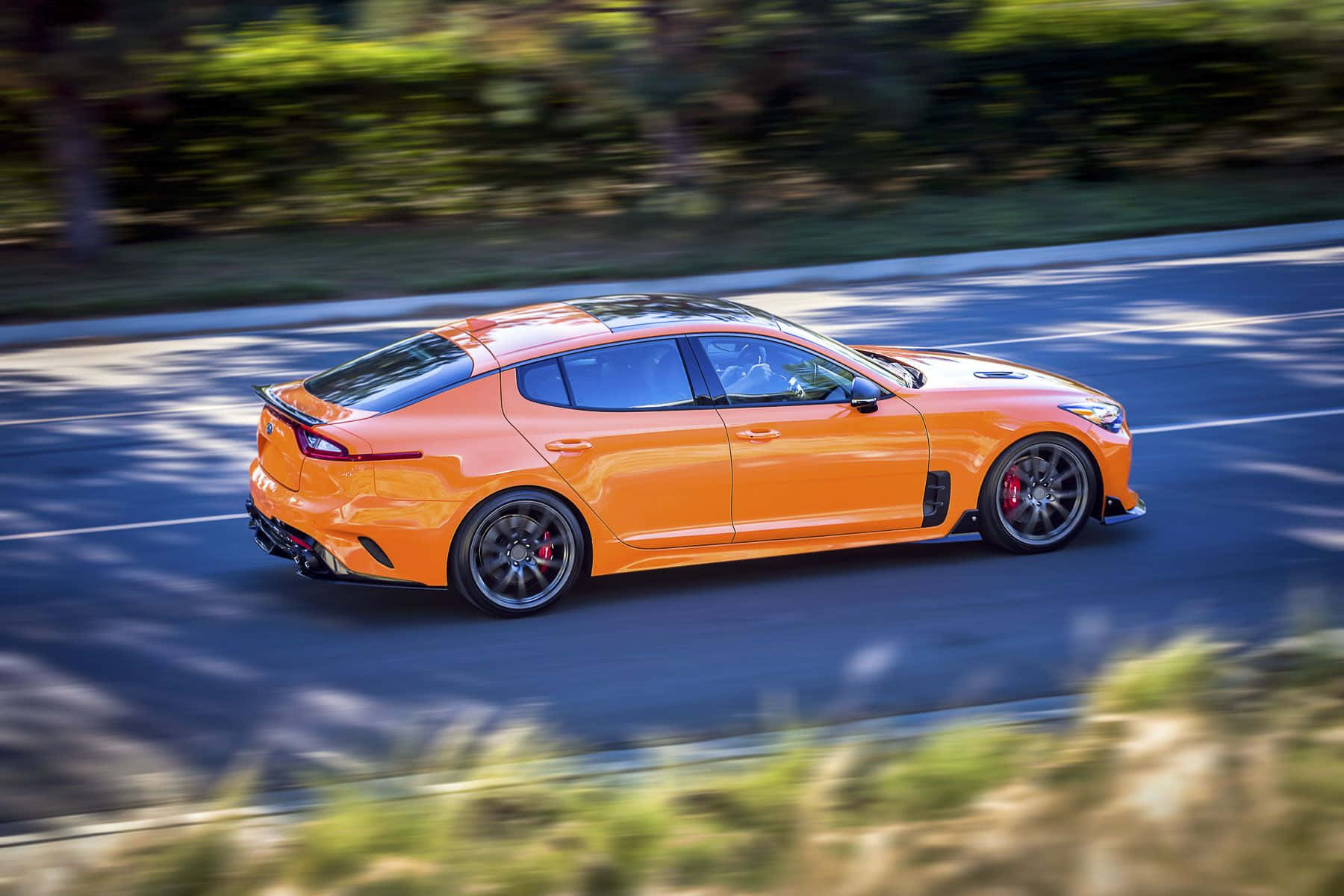 Kia Stinger 桌布 壁紙 — 一輛充滿活力的橙色跑車在陽光明媚的道路上疾馳，周圍的綠色植物模糊移動。
