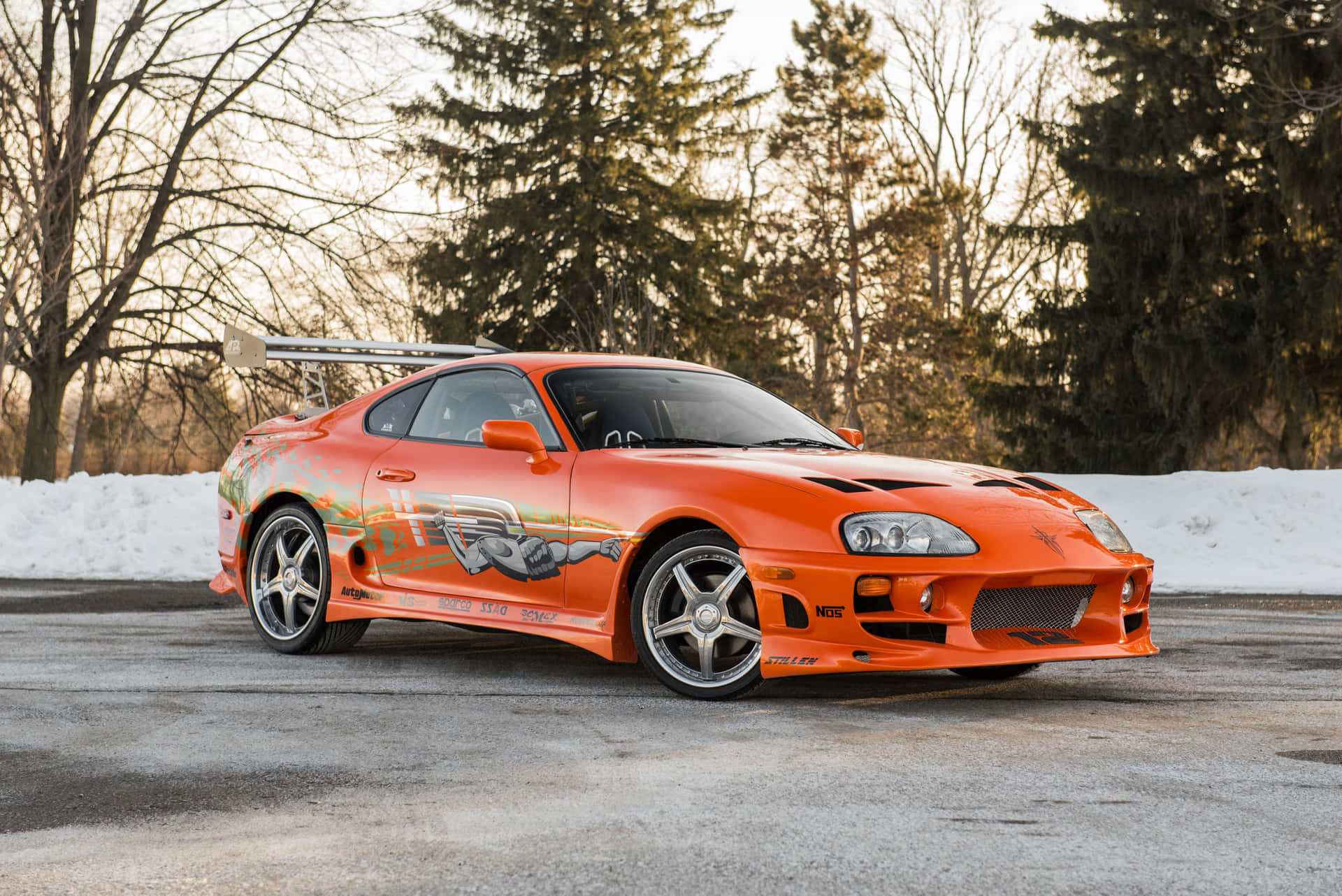 橙色豐田Supra跑車停泊在積雪的道路上，背景有樹木和日落。 — Toyota Supra 壁紙