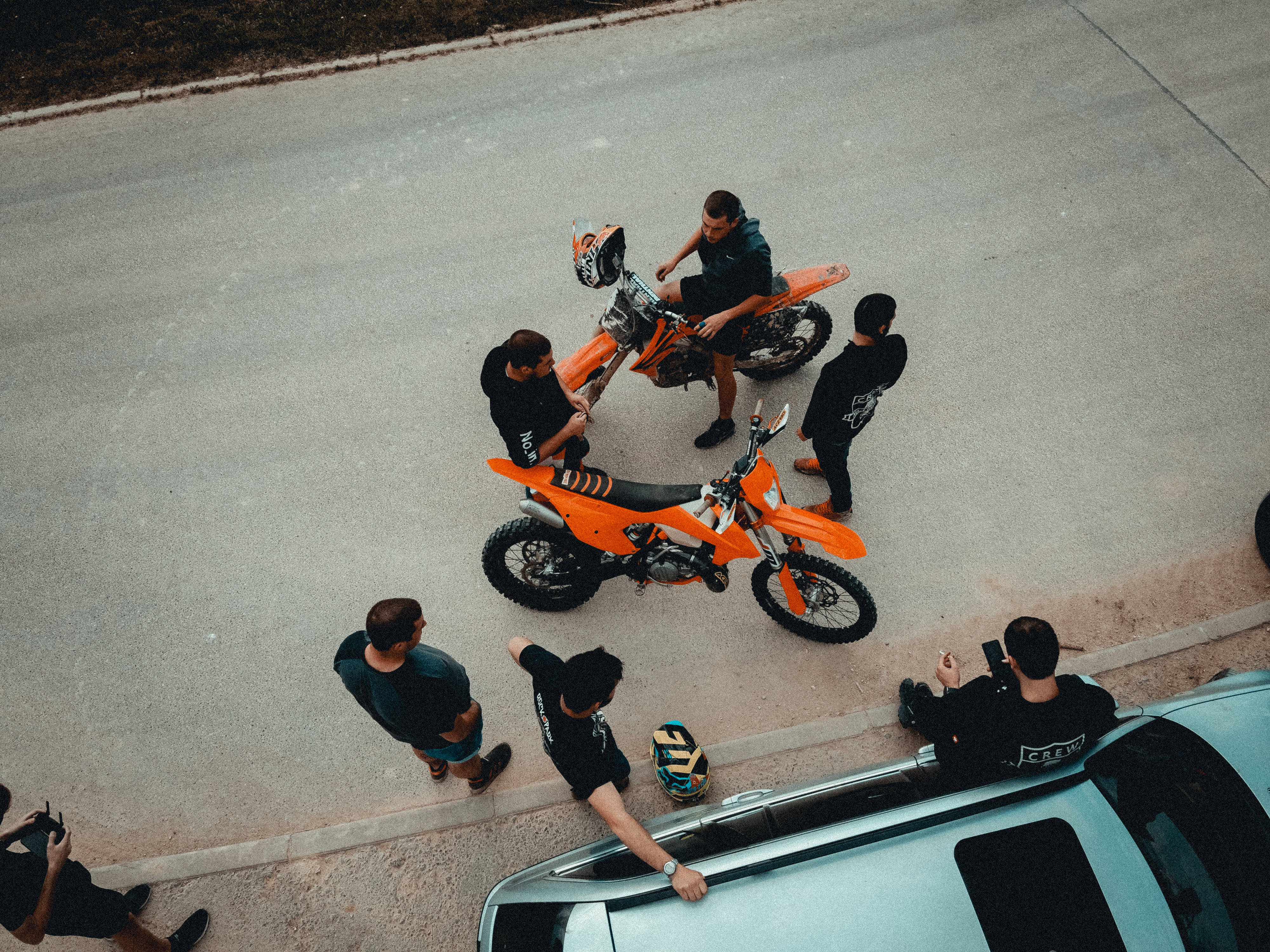 Ktm Bike 桌布 壁紙 — Overhead view of a group of men gathered around two bright orange dirt bikes on a paved road beside a silver car.